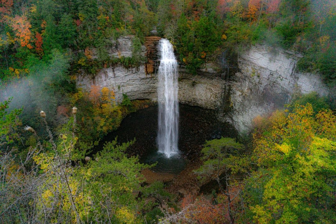 Falls Creek Falls is surrounded by vibrant fall colors and a misty morning haze. The waterfall cascades from a rocky cliff into a dark pool below 13 Scenic Locations to See Autumn Foliage in Tennessee