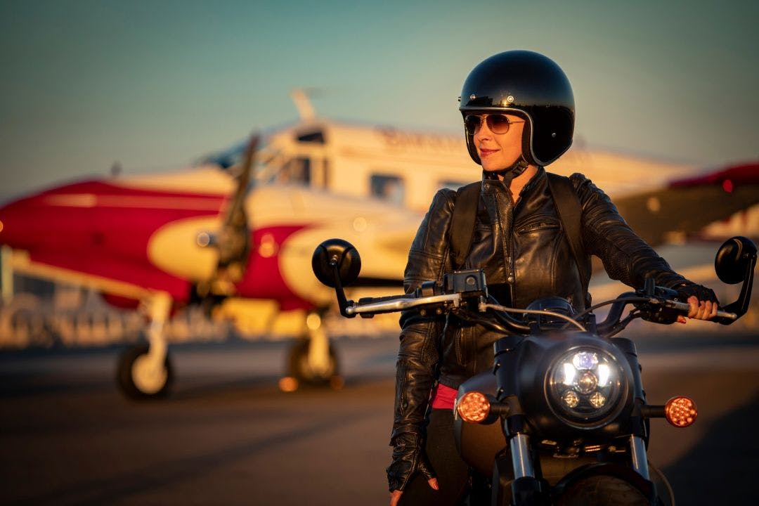 Woman dressed in vintage leather clothes on a motorcycle with an airplane in the background shot at dusk How to Travel With Your Motorcycle Gear on a Plane