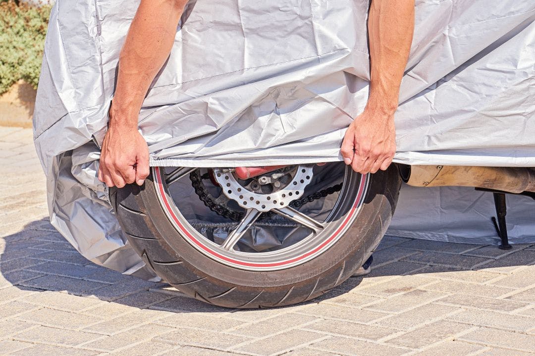 Parked outdoor motorbike covered with protective fabric shield from rain and overheating. Biker covers motorcycle with waterproof protective cover with silver reflective surface Tips for How to Store Your Motorcycle for Winter