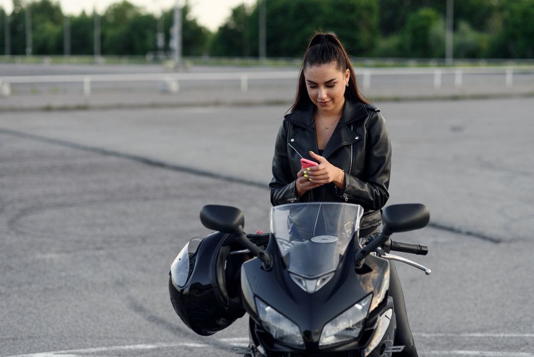 Female biker uses smartphone while sitting on stylish sport motorcycle at outdoors parking. Make Money Renting Your Motorcycle – 2025 Guide