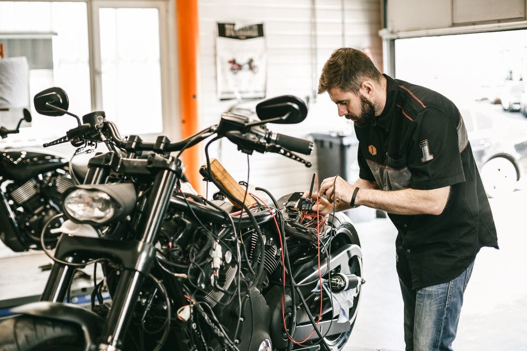 Professional motorcycle mechanic working in bike repair service. Mechanic checking a bike battery level with voltmeter in garage How to Jump Start a Motorcycle Battery – Step-by-Step Guide
