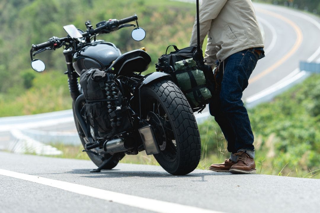 A person stands beside a parked motorcycle on a winding road surrounded by greenery, preparing for a journey or taking a break How to Plan & Prepare for Longer Distance Motorcycle Trips