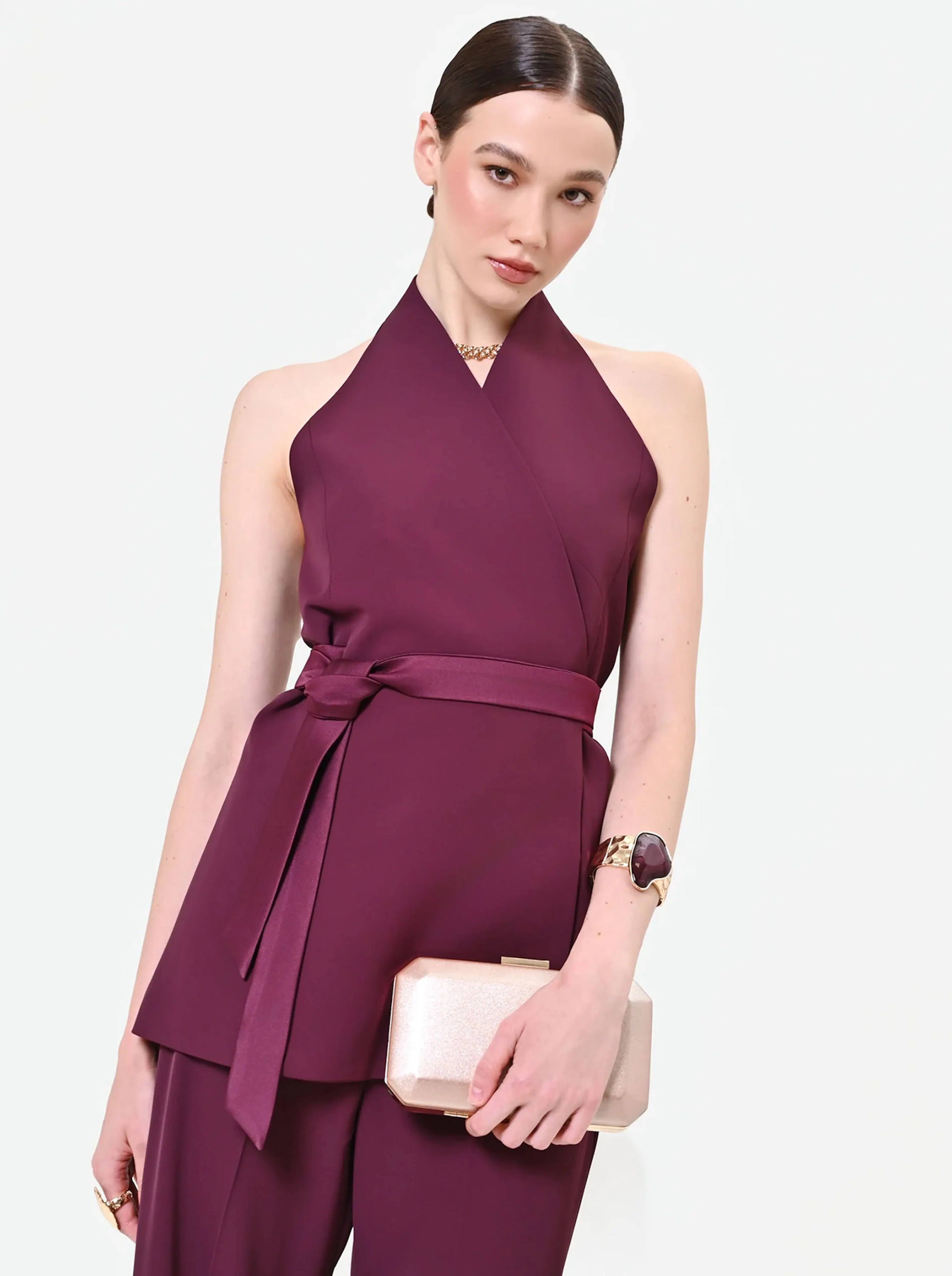 Woman in a sleeveless, deep burgundy halter top and matching pants, holding a pale pink clutch. She exudes elegance and confidence against a plain white background.