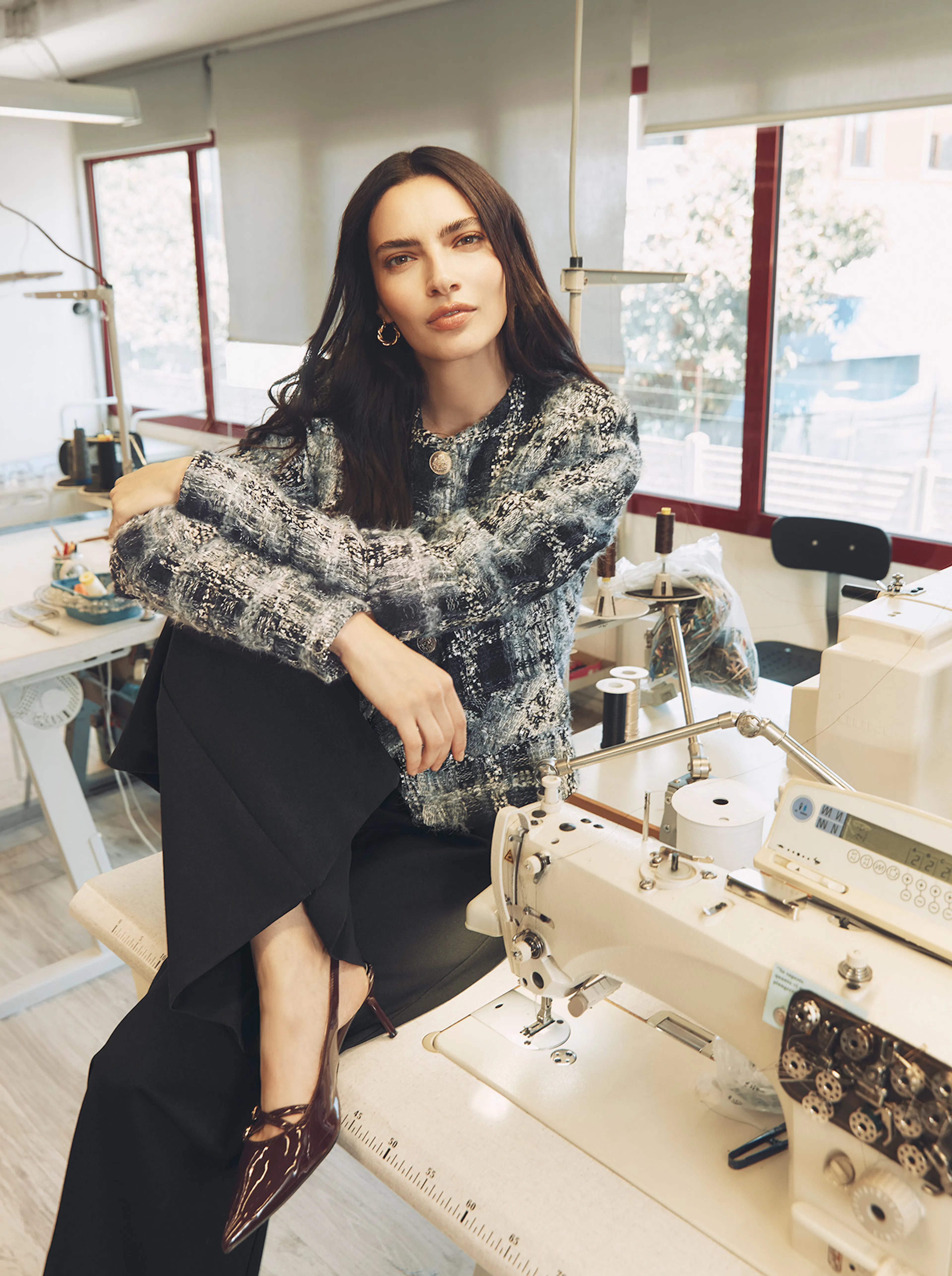 A brunette model sits on the edge of a workstation next to a sewing machine in a tailoring studio. She is wearing a textured blue, black, and white tweed jacket with gold buttons, paired with black trousers and dark burgundy pointed-toe slingback heels.