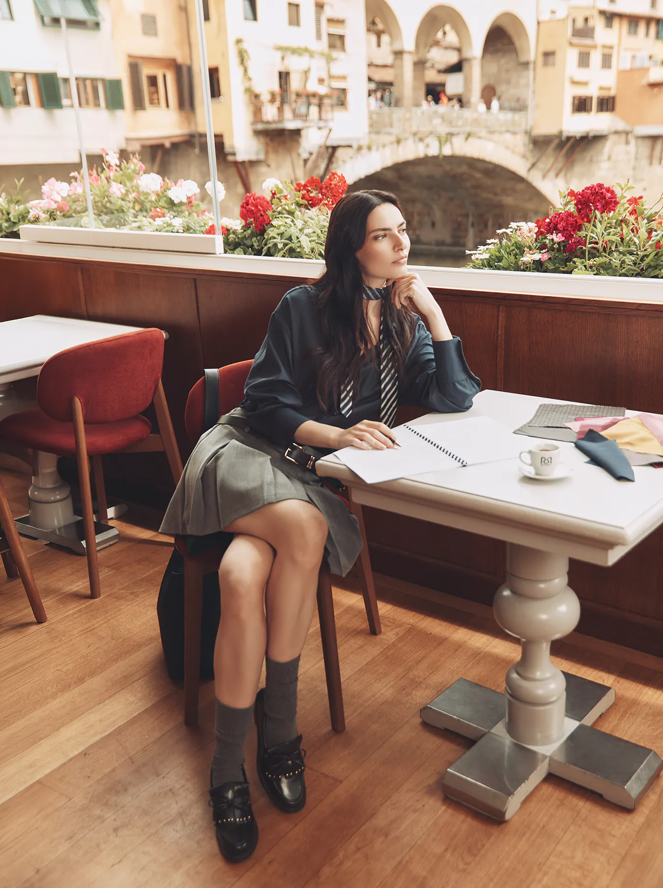 A brunette model sits at a small cafe table by a large window overlooking an Italian river and bridge. She is dressed in a dark blue blouse with a striped tie, a grey pleated mini skirt, grey socks, and black loafers. She holds a pen over an open notebook, looking thoughtfully out the window with fabric swatches on the table.