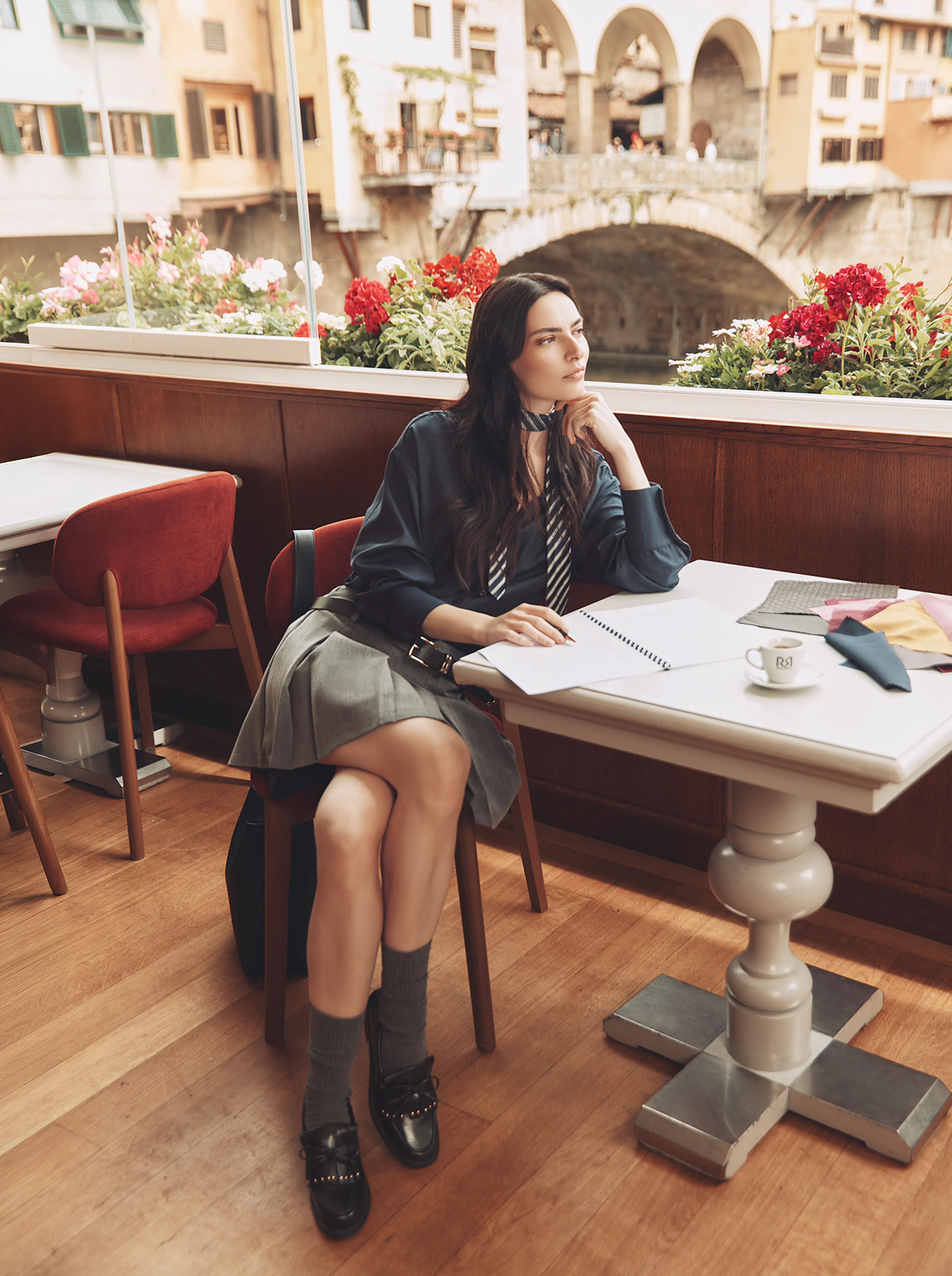 A brunette model sits at a small cafe table by a large window overlooking an Italian river and bridge. She is dressed in a dark blue blouse with a striped tie, a grey pleated mini skirt, grey socks, and black loafers. She holds a pen over an open notebook, looking thoughtfully out the window with fabric swatches on the table.