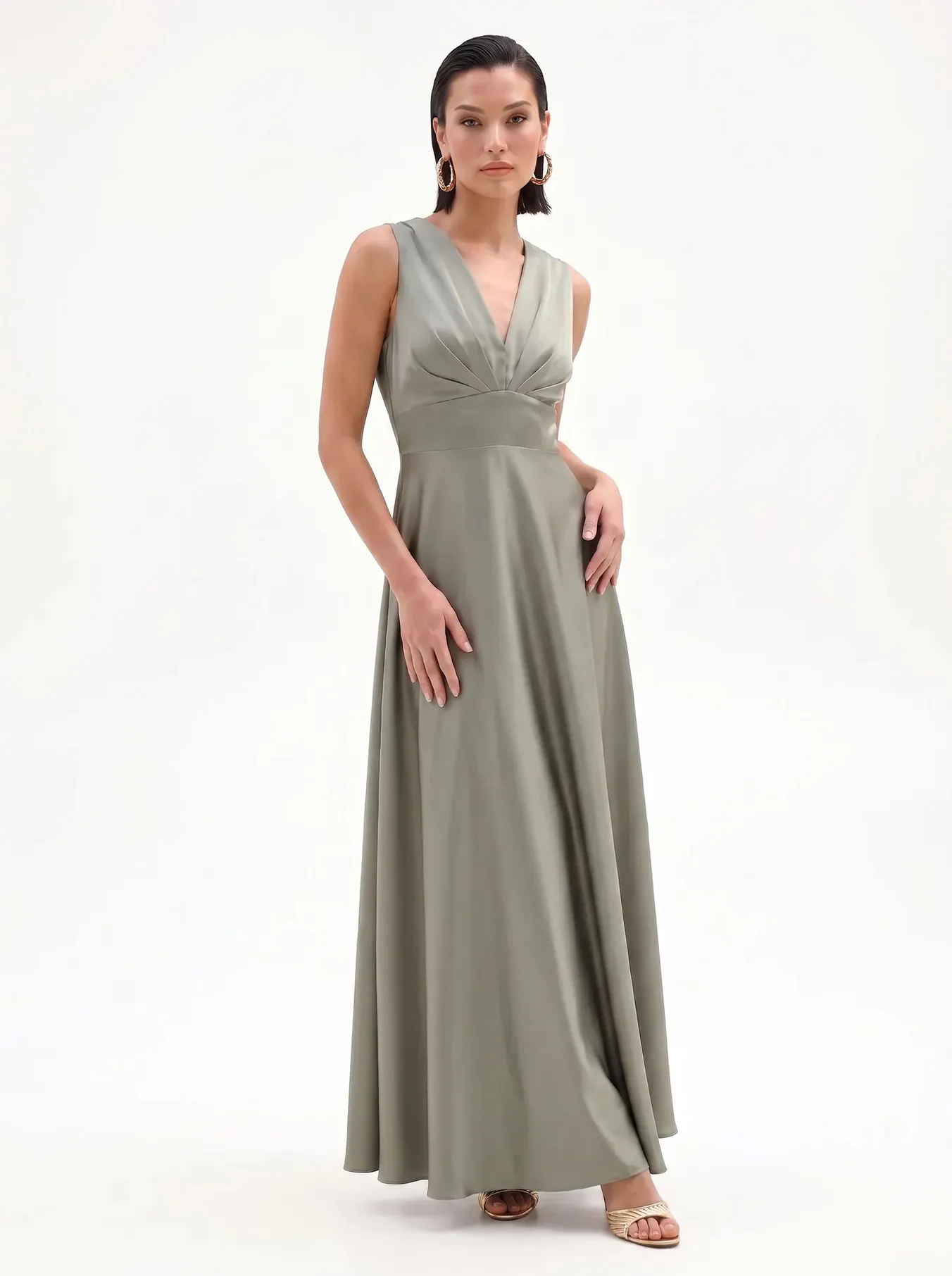 A woman in a flowing, sage green gown stands against a plain background. She holds a beige clutch and wears elegant heels, exuding a poised and refined demeanor.
