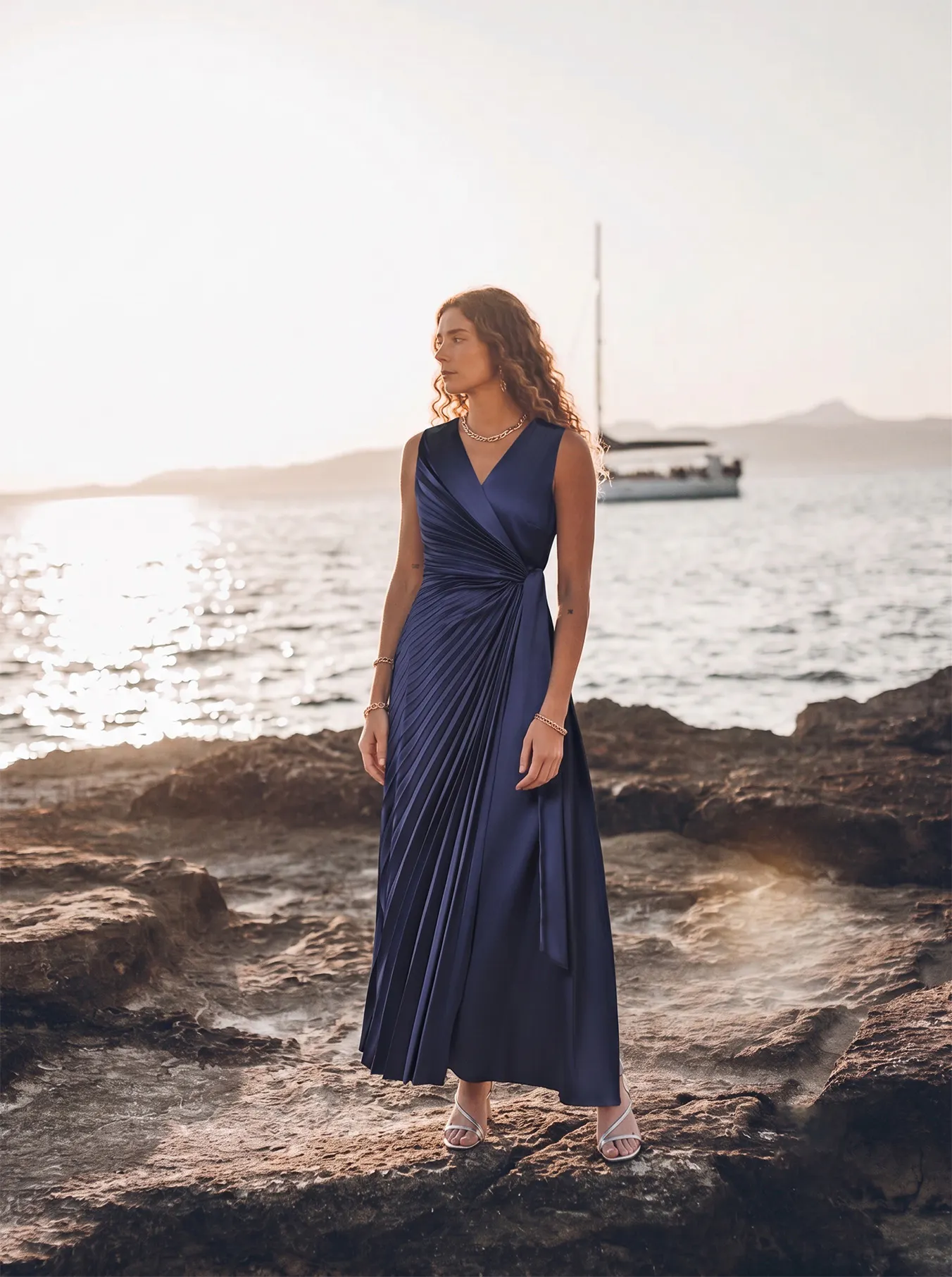 A woman in a blue dress stands on rocky beach at sunset, gazing towards the sea. A sailboat is visible in the background, evoking a serene, elegant atmosphere.