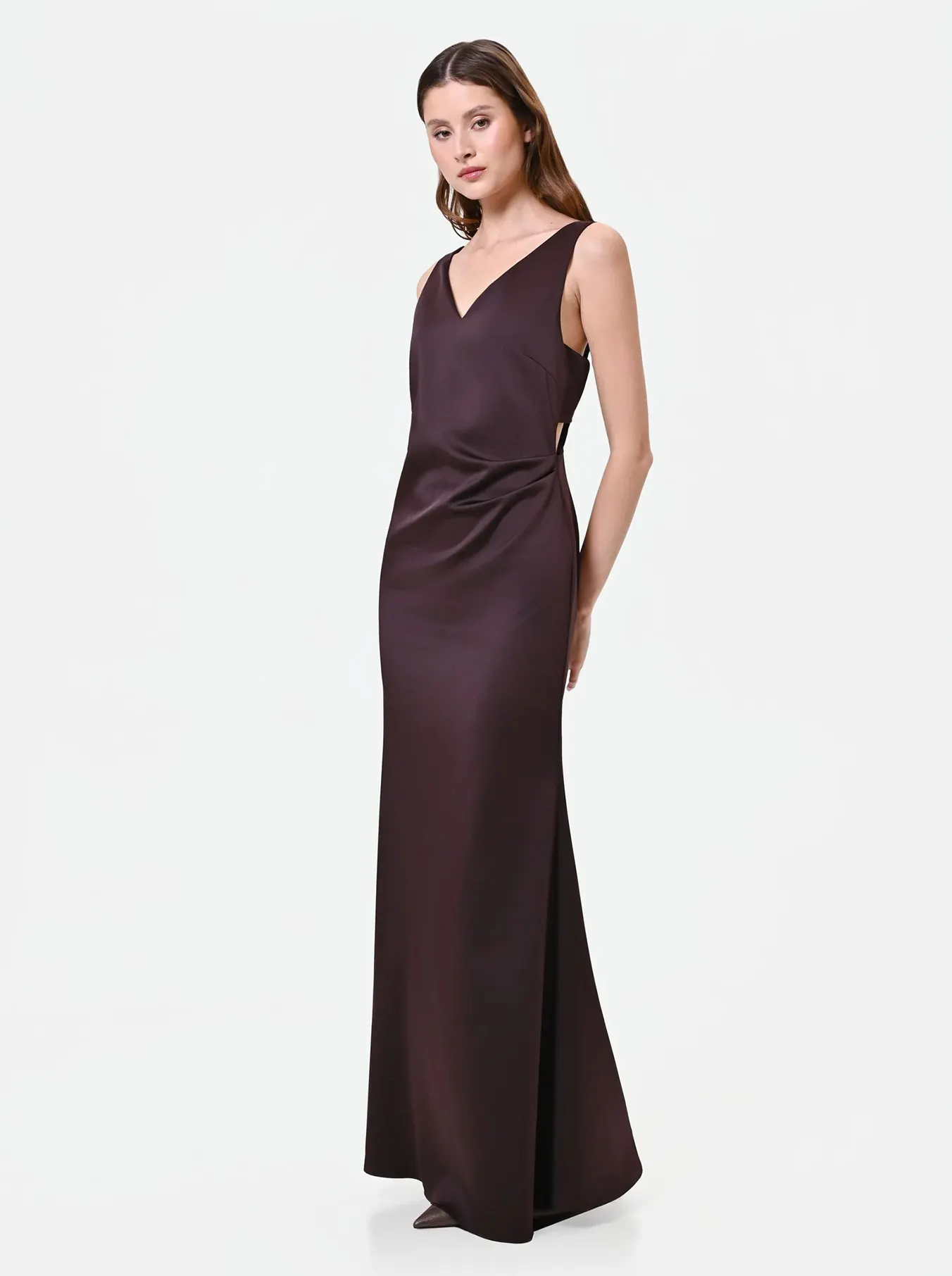 Elegant woman in a sleeveless, floor-length dark brown dress with a V-neckline. She poses with a serene expression against a white background.