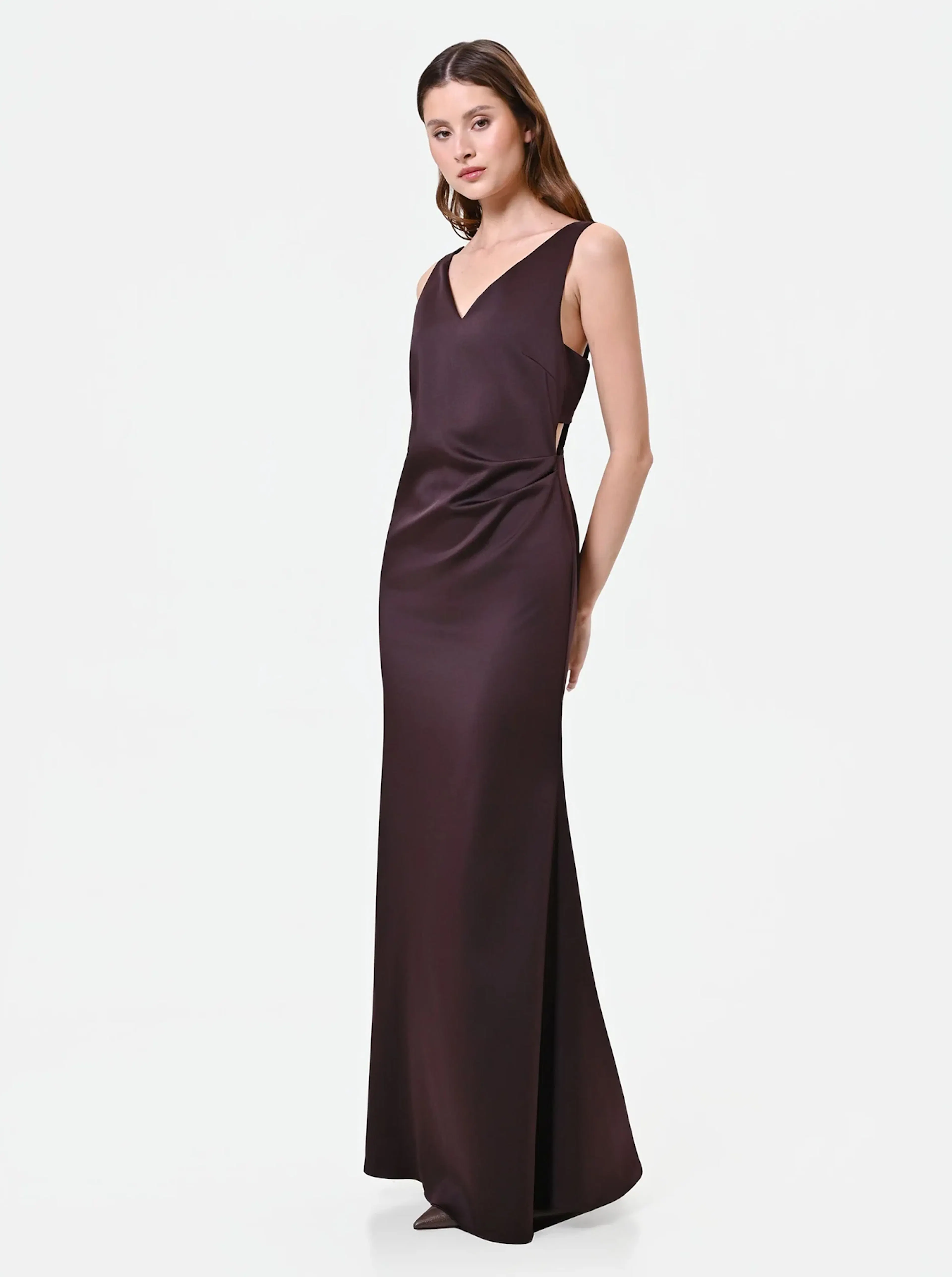 Elegant woman in a sleeveless, floor-length dark brown dress with a V-neckline. She poses with a serene expression against a white background.
