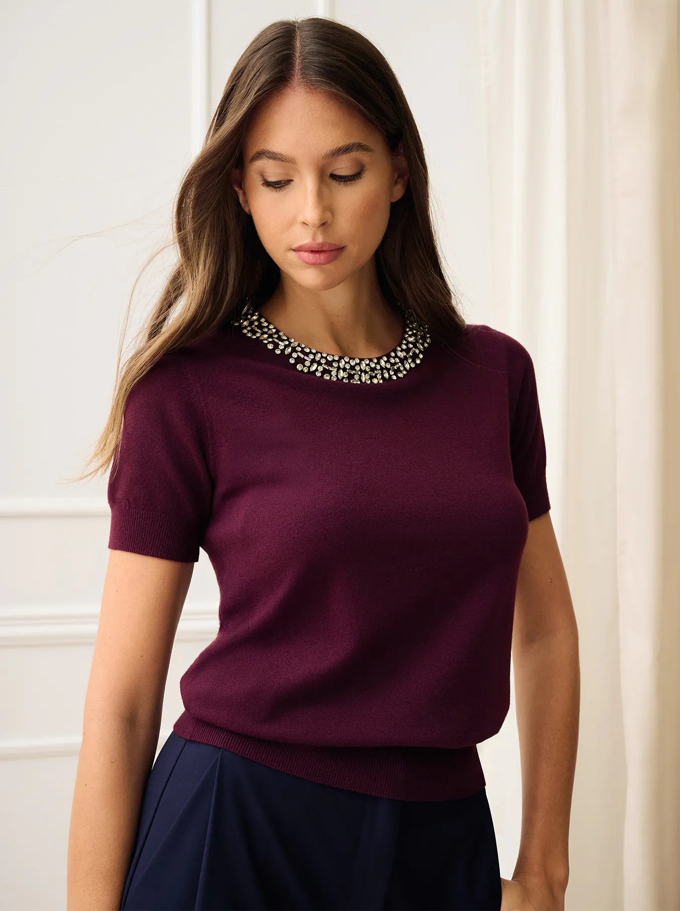 A woman models a plum short-sleeve top with a sparkling jeweled neckline and navy skirt, looking down against a bright, elegant interior.
