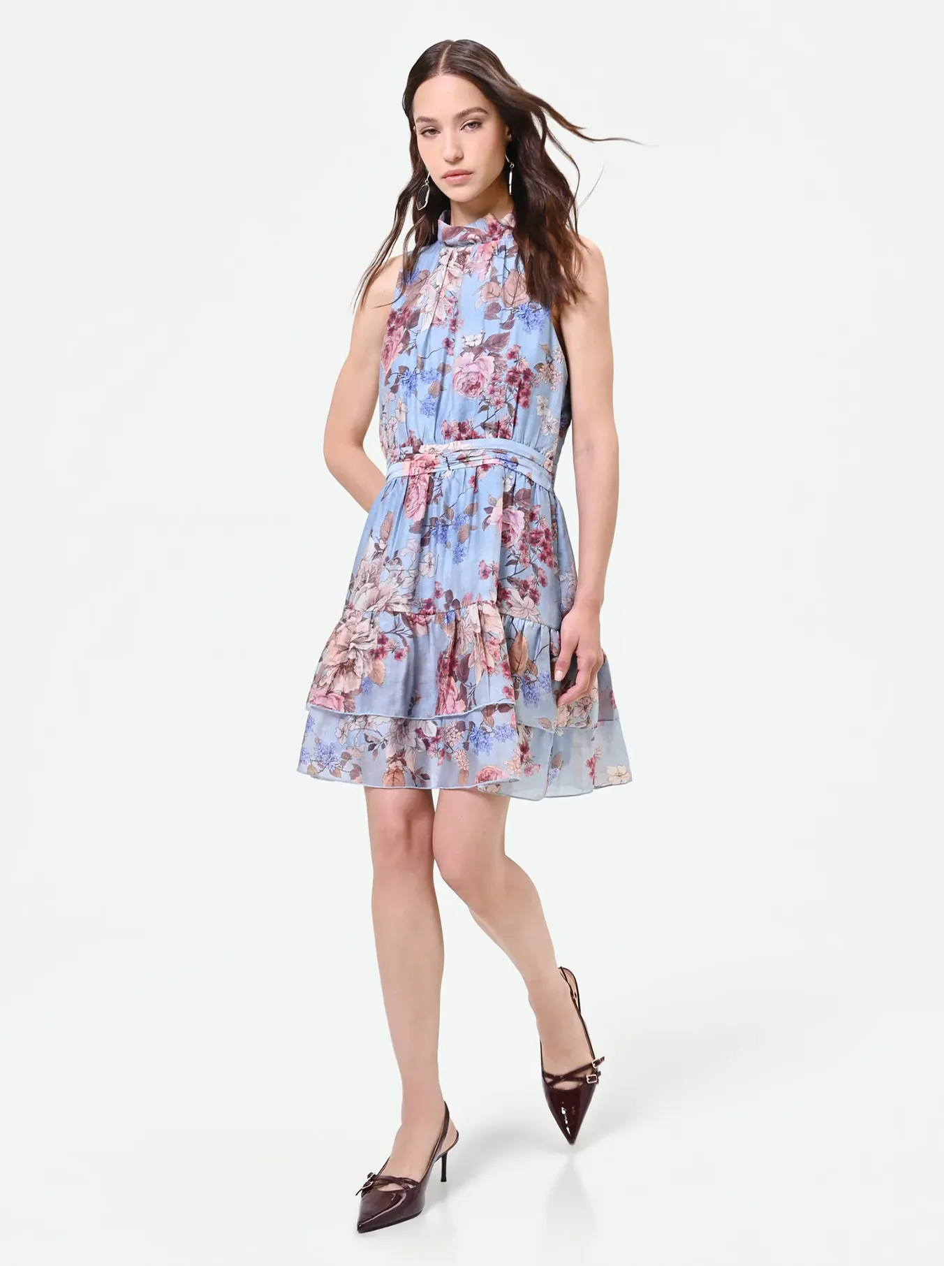 A woman in a blue floral-print sleeveless dress and dark heels poses confidently against a plain white backdrop, exuding elegance and grace.