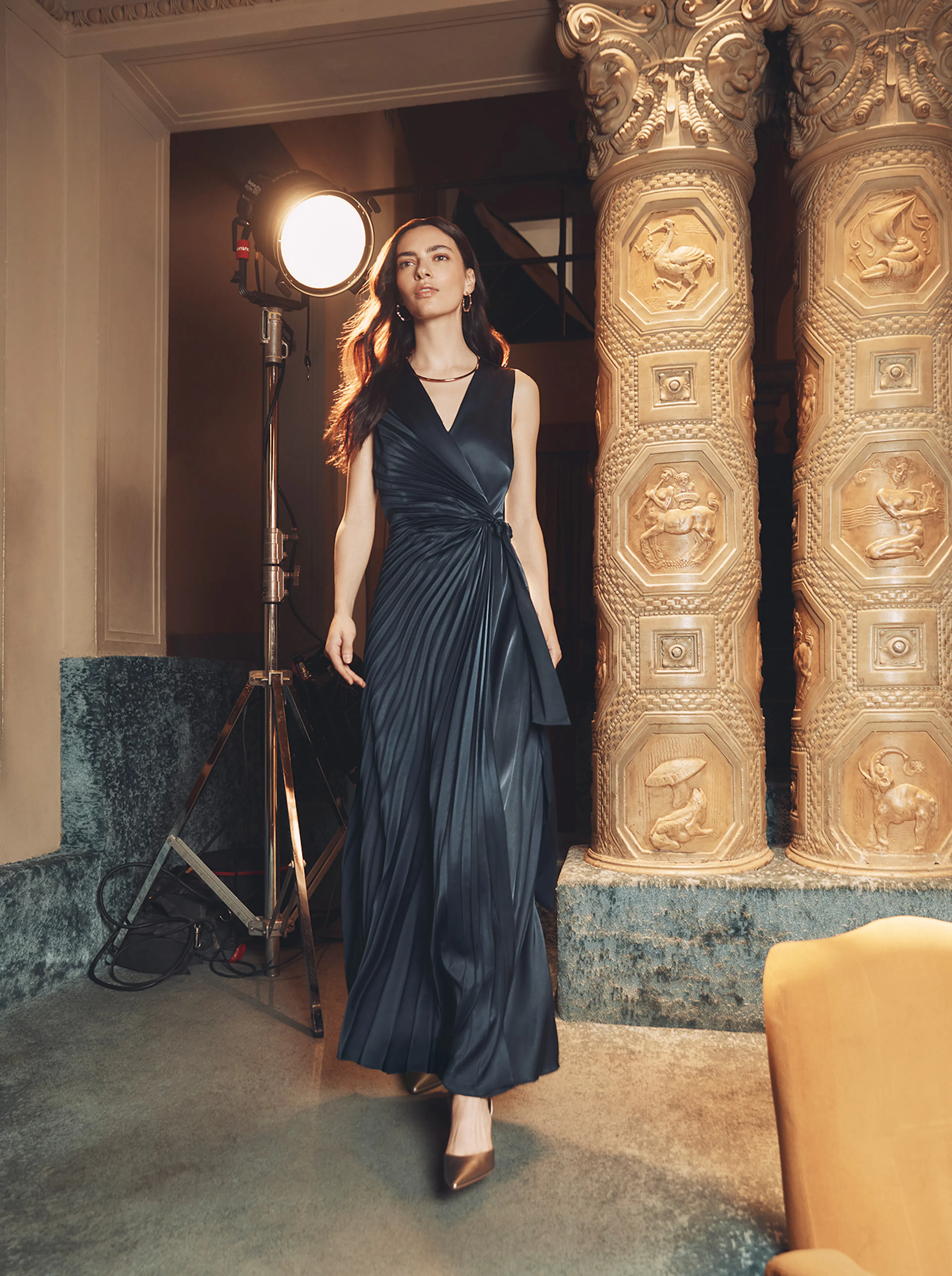 A brunette model walks confidently past ornate, carved stone pillars in a grand room. She is wearing a dark, sleeveless pleated wrap maxi dress and metallic pointed-toe heels. A large studio photography light stands to her left.