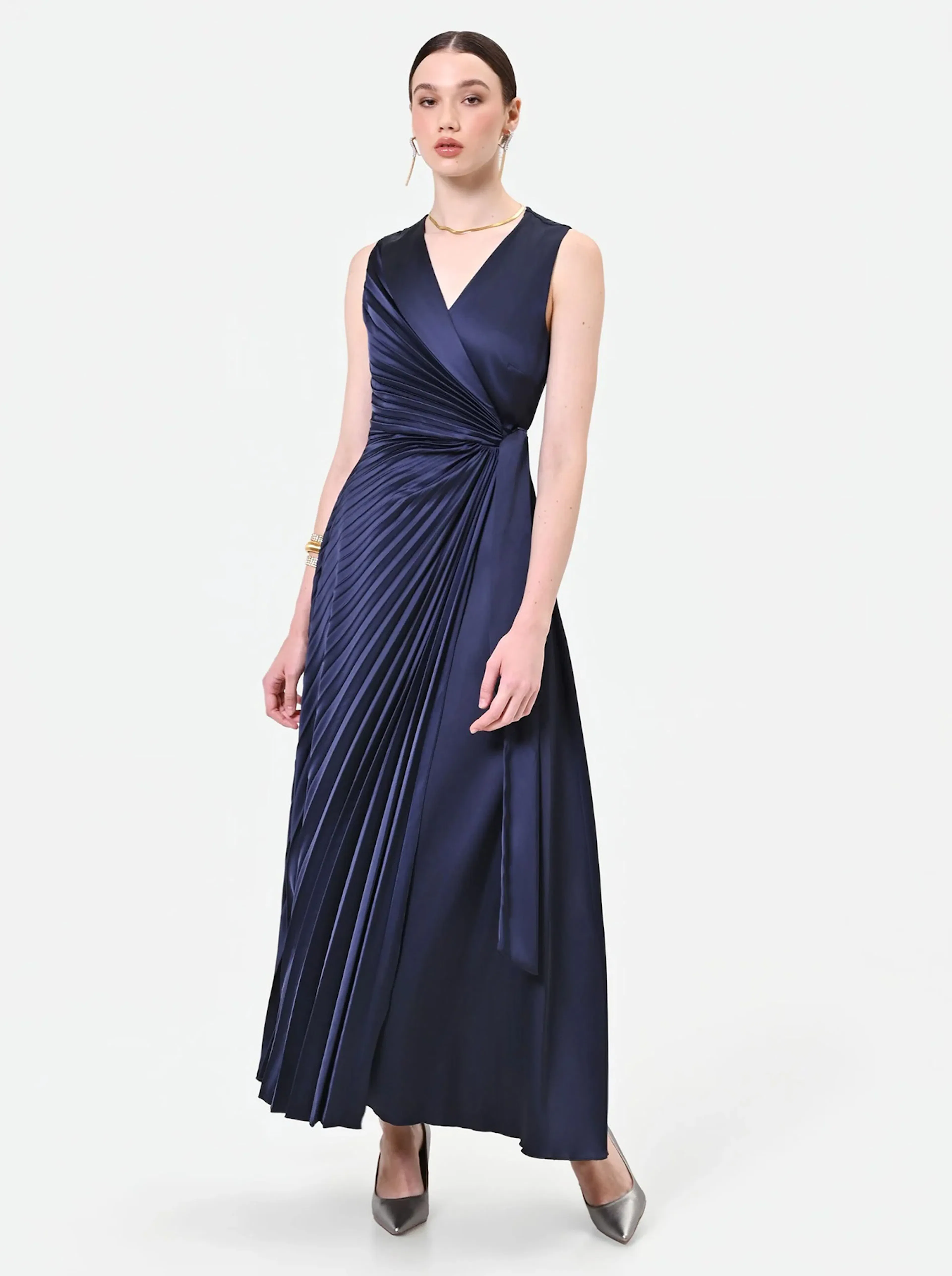 A woman in a navy blue pleated gown stands confidently against a white background. The dress features a wrap design and metallic heels. The tone is elegant.