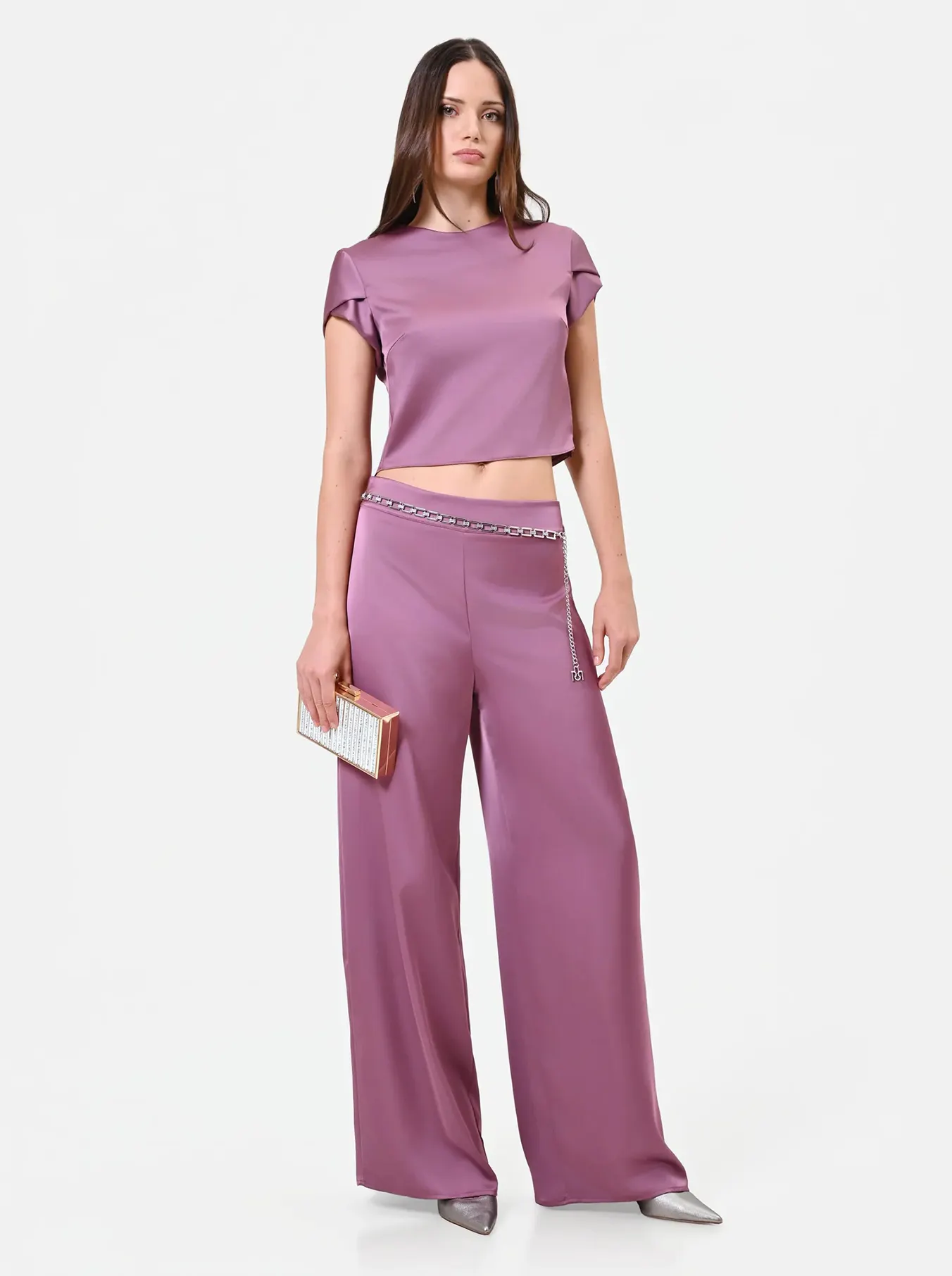 Woman in a purple satin outfit with a chain belt holds a gold clutch. Her expression is neutral, exuding elegance and modernity against a white background.