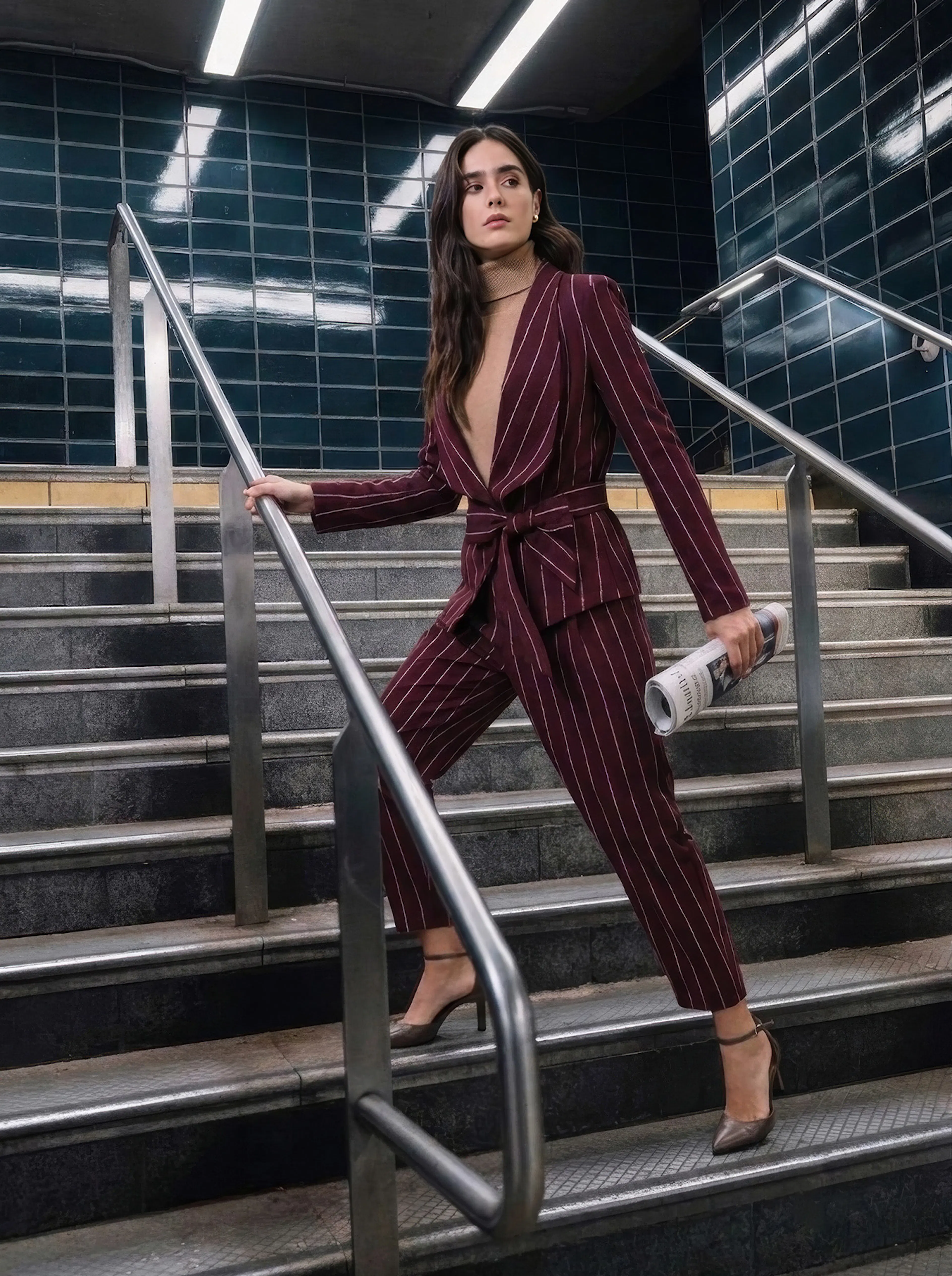 Fashionable woman in a burgundy pinstriped suit descends a subway staircase, holding the rail and a rolled magazine, poised and confident.