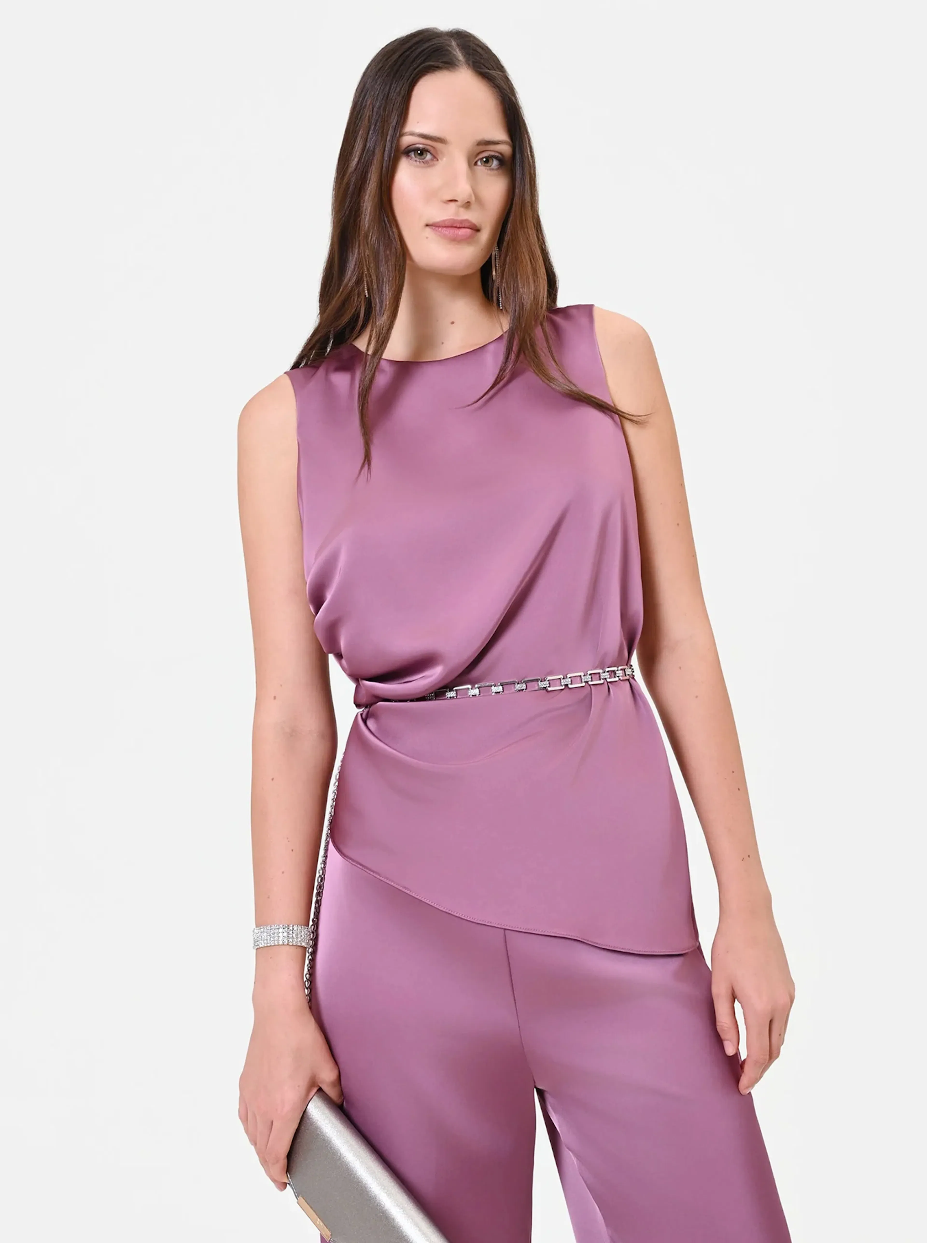 Woman in a sleeveless mauve blouse with a silver chain belt and matching clutch. Her expression is confident and elegant against a plain background.