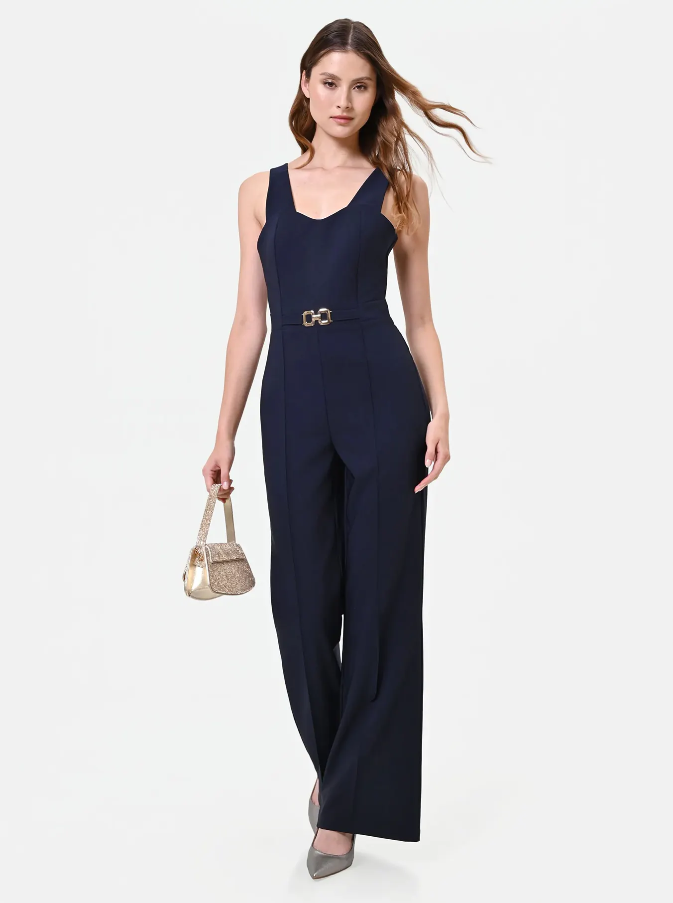 Woman in a navy jumpsuit with a fitted waist and wide legs, holding a gold handbag. Her hair flows, and her expression is confident and elegant.