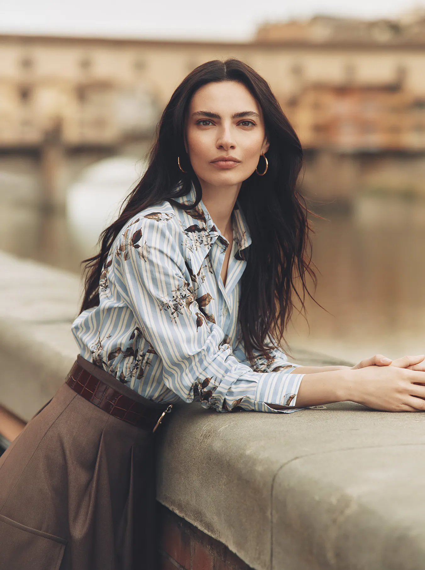 A close-up portrait of a brunette model leaning against a stone wall overlooking a river, with a historic bridge reminiscent of Florence's Ponte Vecchio in the softly blurred background. She is wearing a light blue and white striped shirt with floral print details, tucked into a brown skirt with a brown croc-embossed belt.