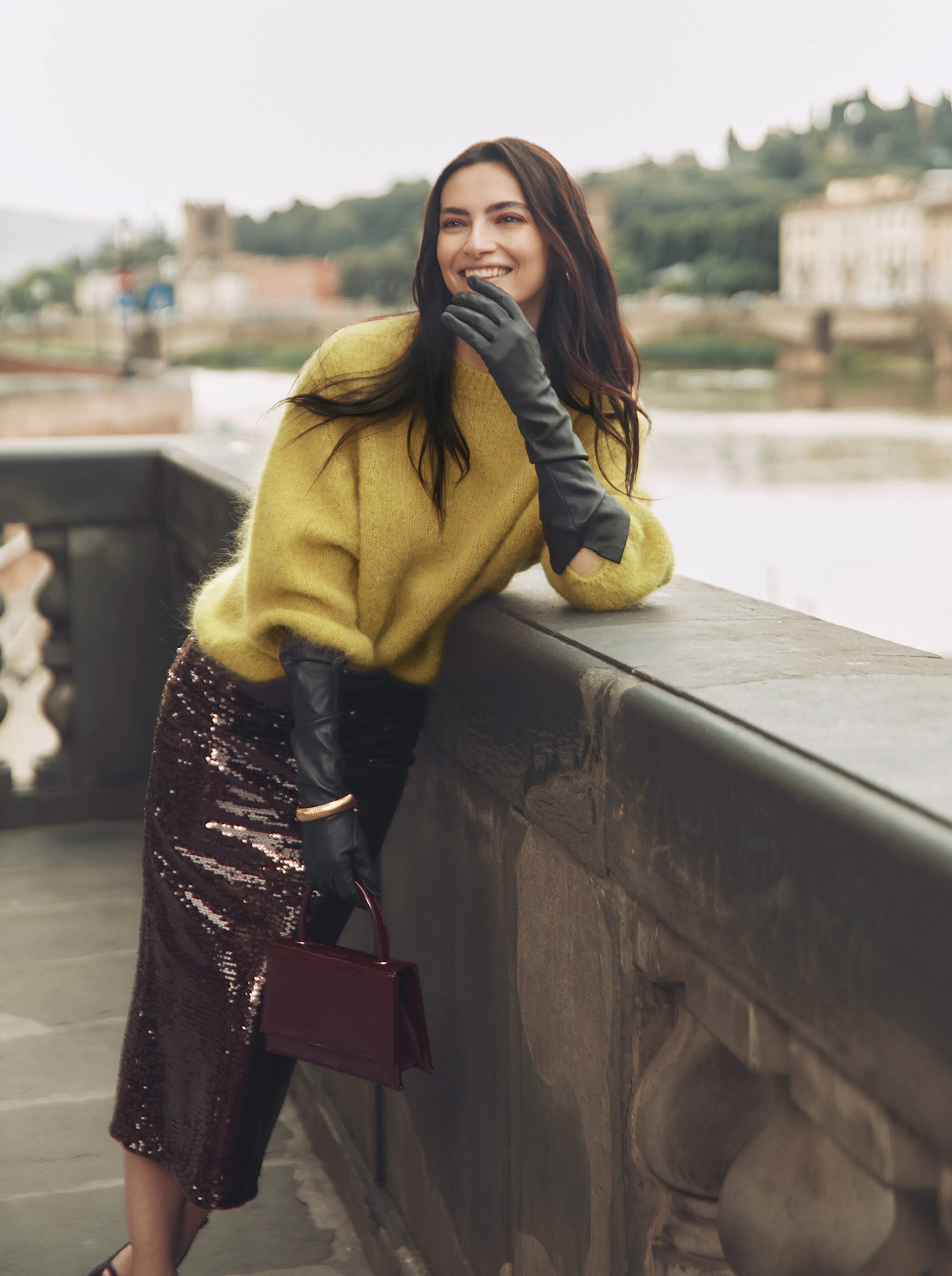 Woman in a yellow sweater, black gloves, and sequined skirt leans on a stone bridge railing, holding a small handbag, smiling outdoors.