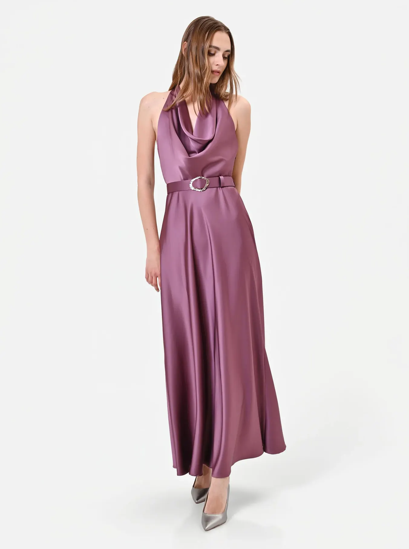 A woman wearing a silky, mauve halter-neck gown with a draped neckline and a matching belt stands against a plain white background. She looks down, exuding elegance.