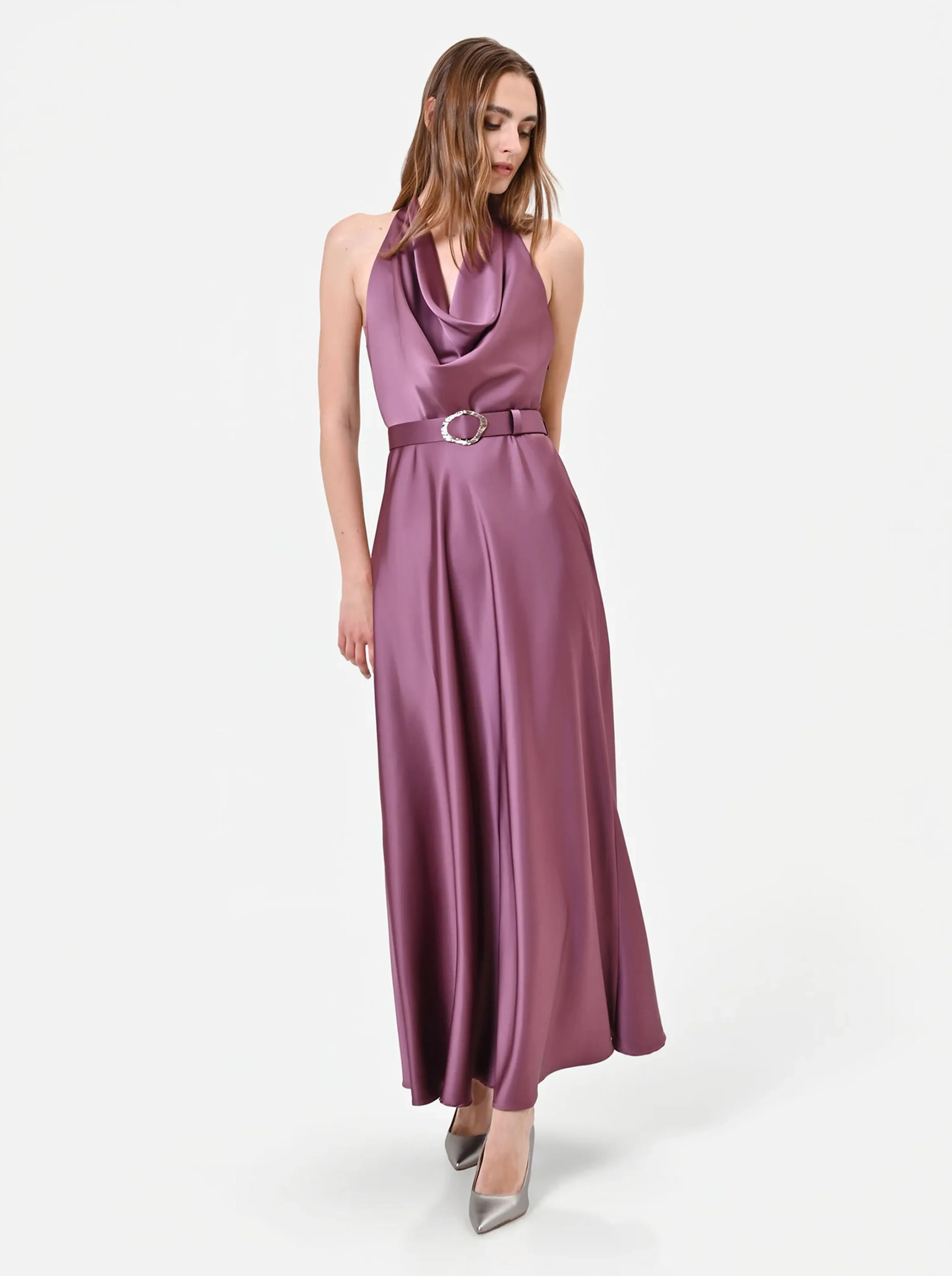 A woman wearing a silky, mauve halter-neck gown with a draped neckline and a matching belt stands against a plain white background. She looks down, exuding elegance.