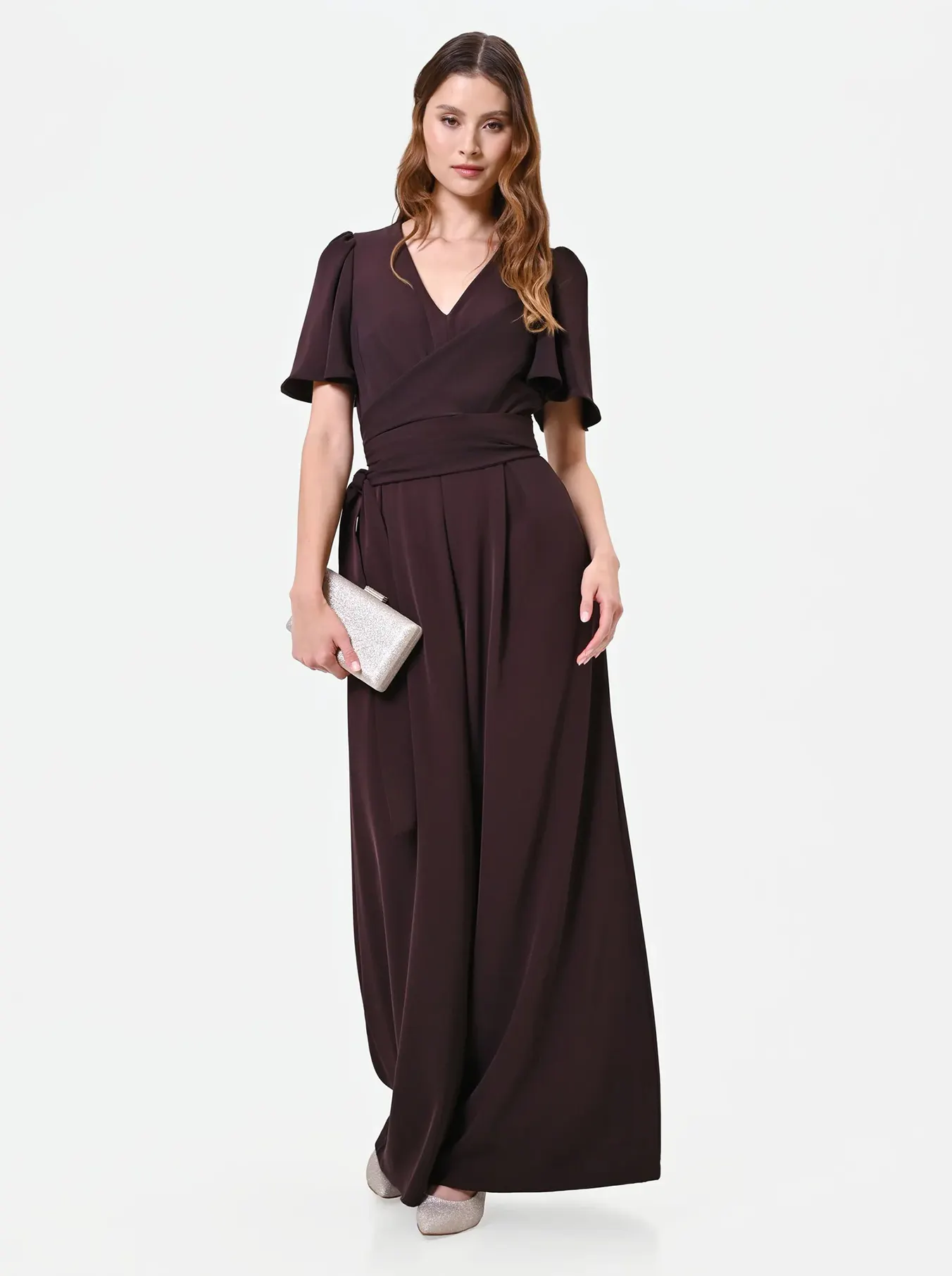 Woman in an elegant, dark brown floor-length jumpsuit holds a silver clutch. She stands confidently against a plain white background, exuding sophistication.