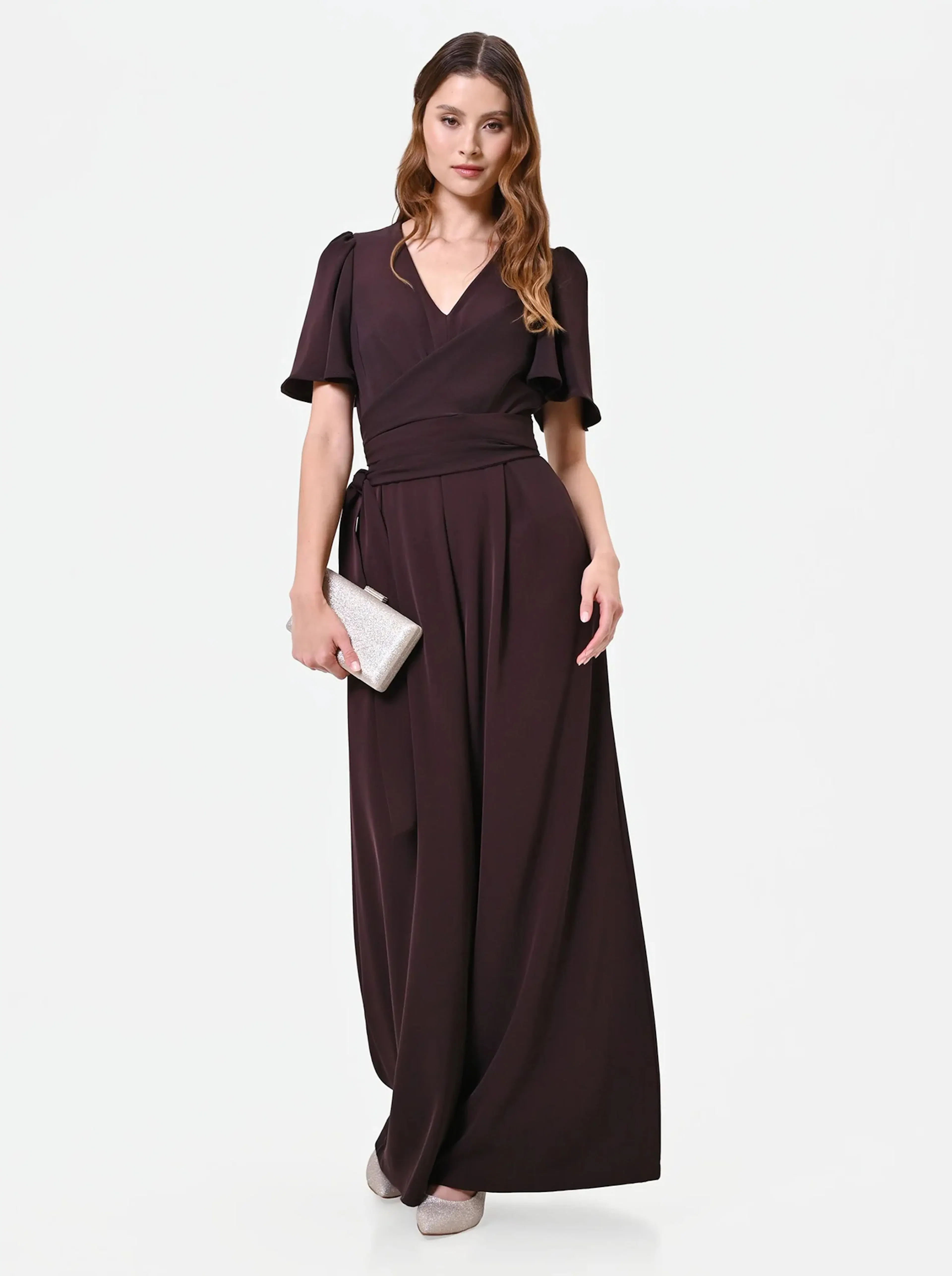 Woman in an elegant, dark brown floor-length jumpsuit holds a silver clutch. She stands confidently against a plain white background, exuding sophistication.