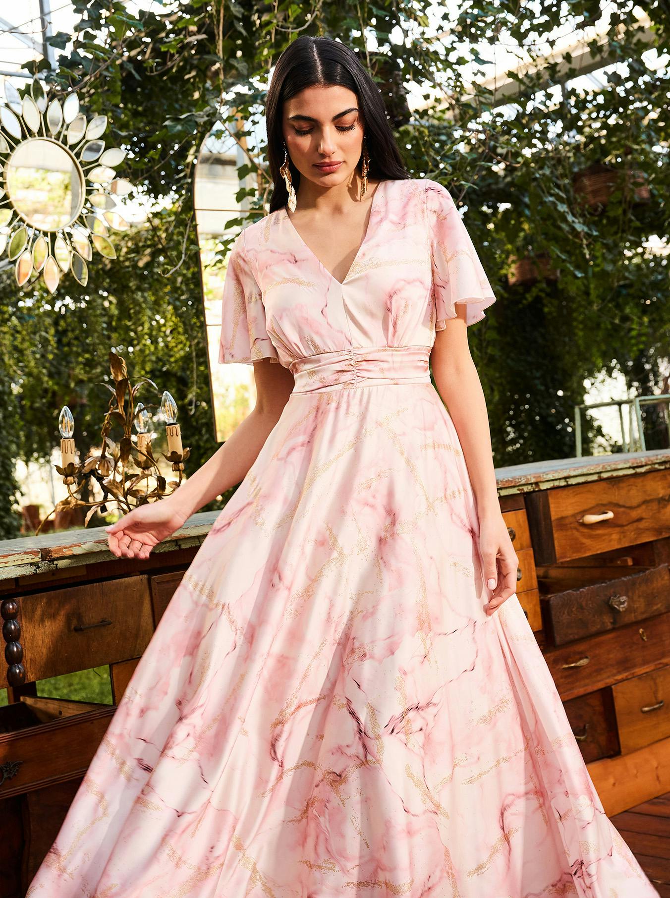 Woman in a pink marble-patterned dress with flutter sleeves stands near a wooden counter, surrounded by greenery and a chandelier.