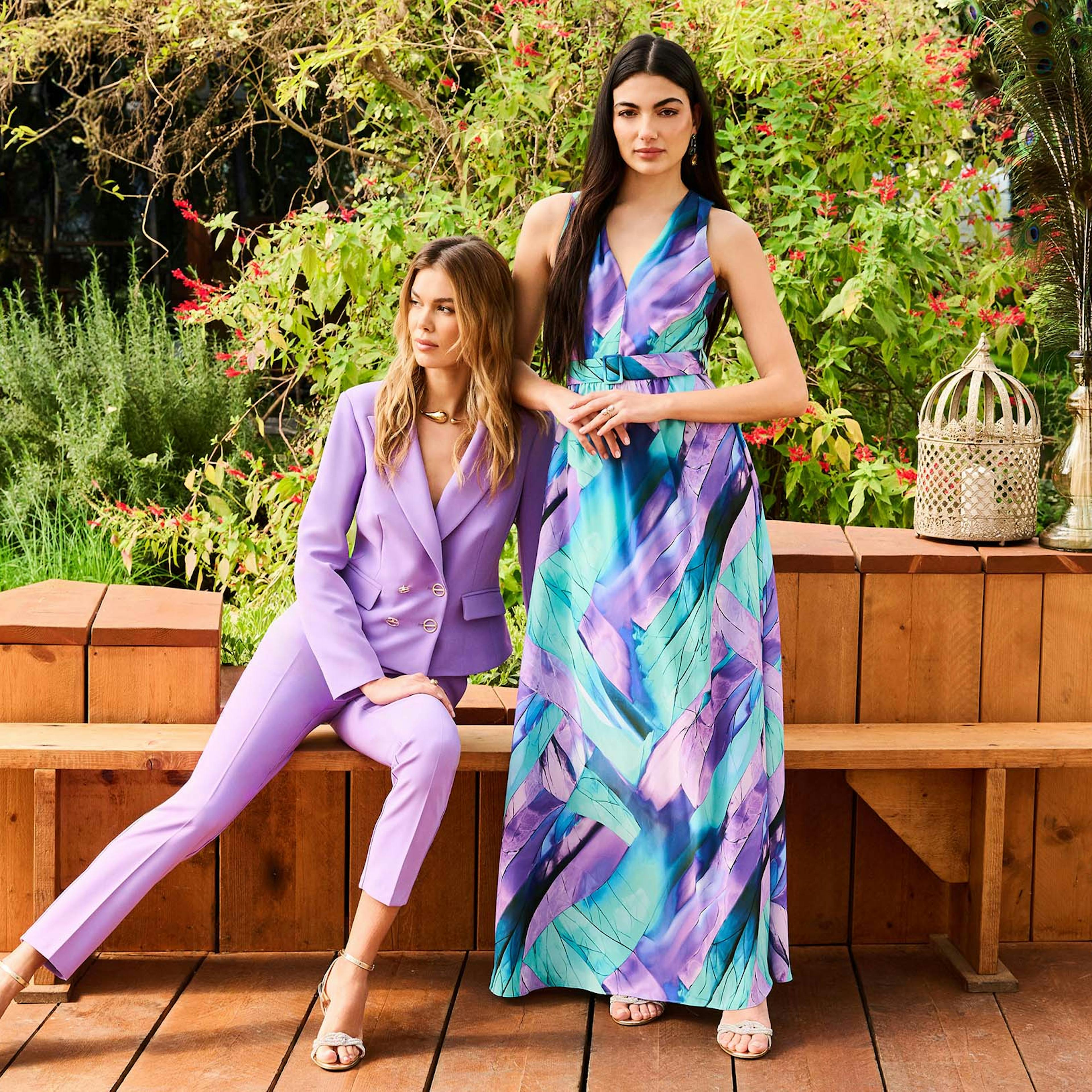 Two women wearing purple and blue outfits sit together on a wooden bench