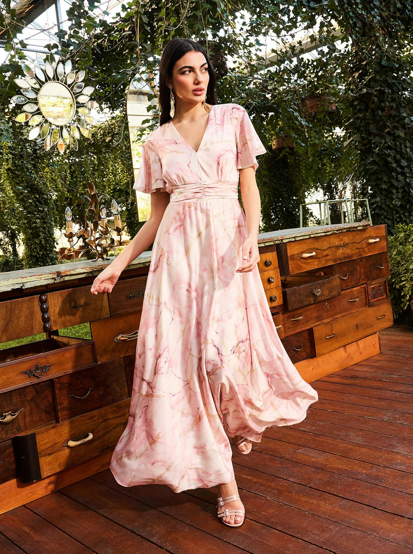 A woman in a flowing pink marble-patterned dress stands on a wooden deck surrounded by lush greenery. She gazes thoughtfully, conveying elegance.