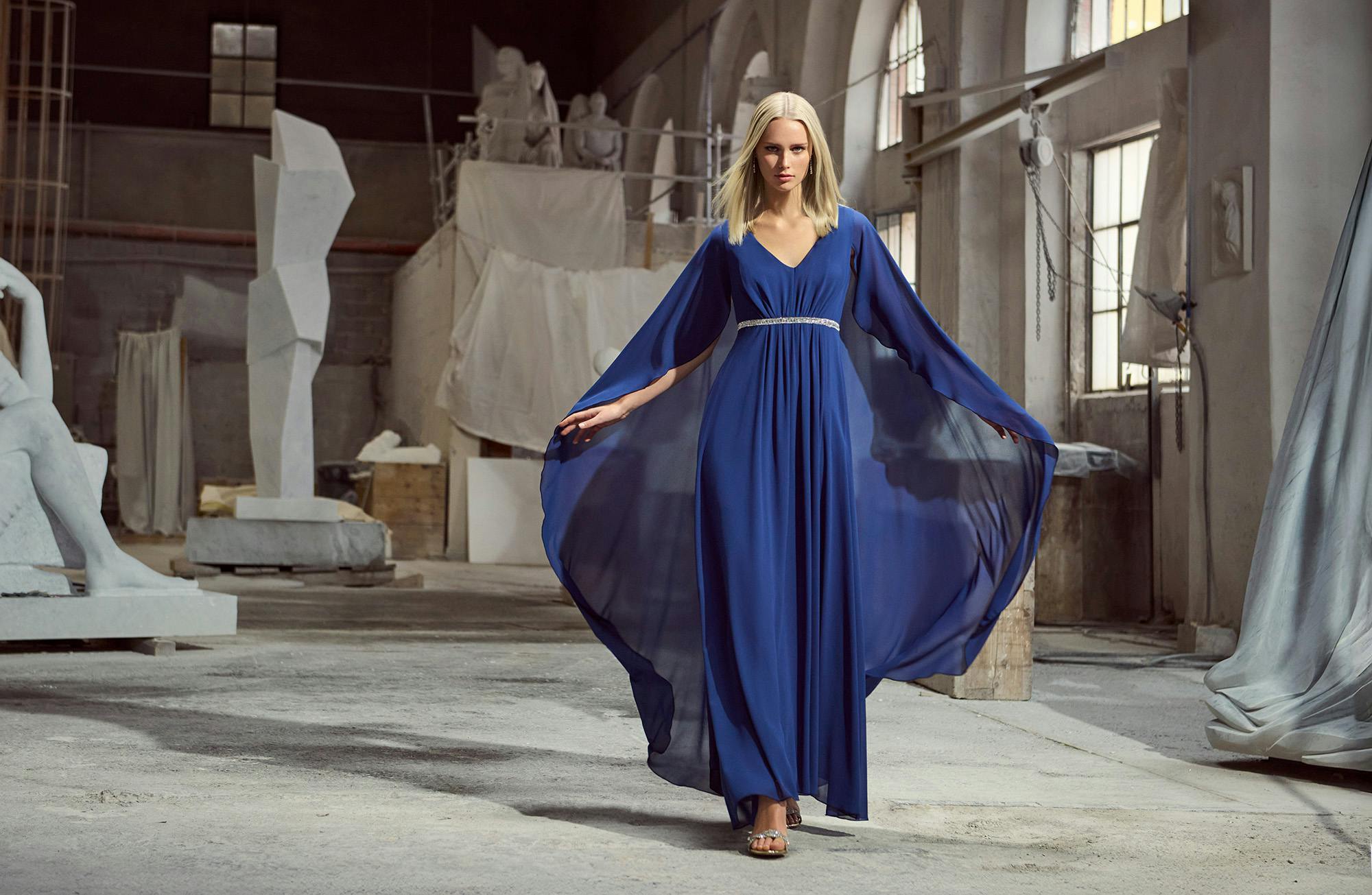A woman in a blue dress stands in front of a large, abstract sculpture in an outdoor setting.
