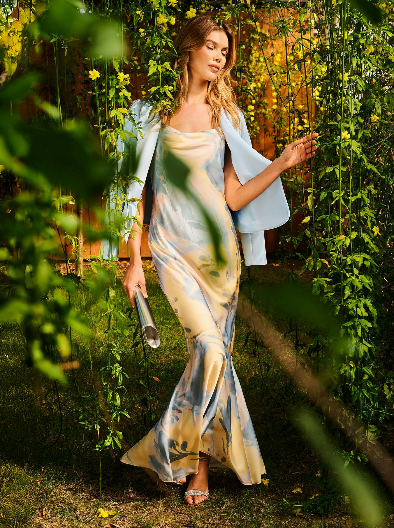A woman in a flowing, pastel-colored dress and light blue shawl walks gracefully through a garden adorned with vibrant yellow flowers.
