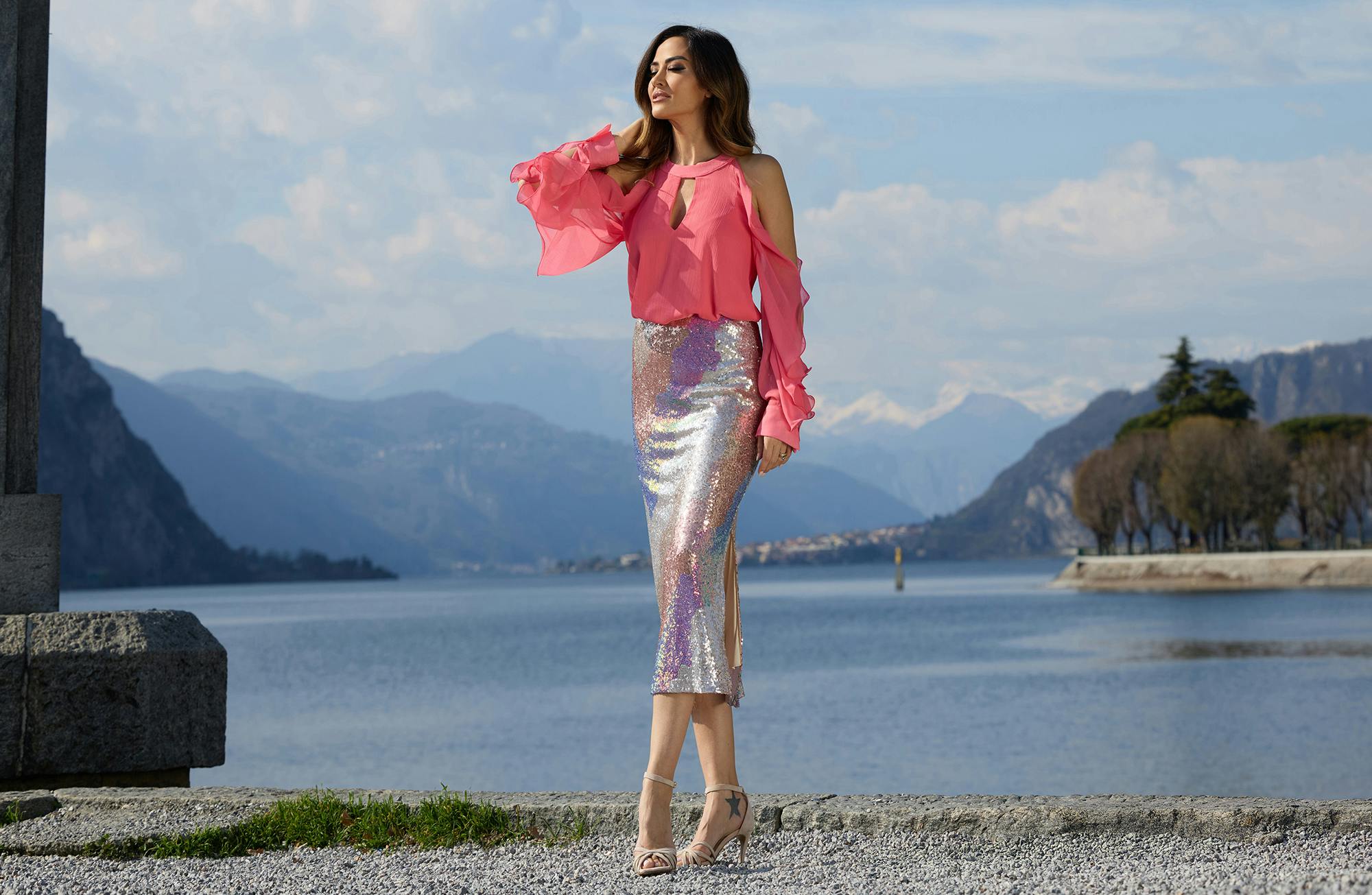 Giorgia Palmas in a pink blouse and shimmering skirt stands by a lake, with mountains in the background.