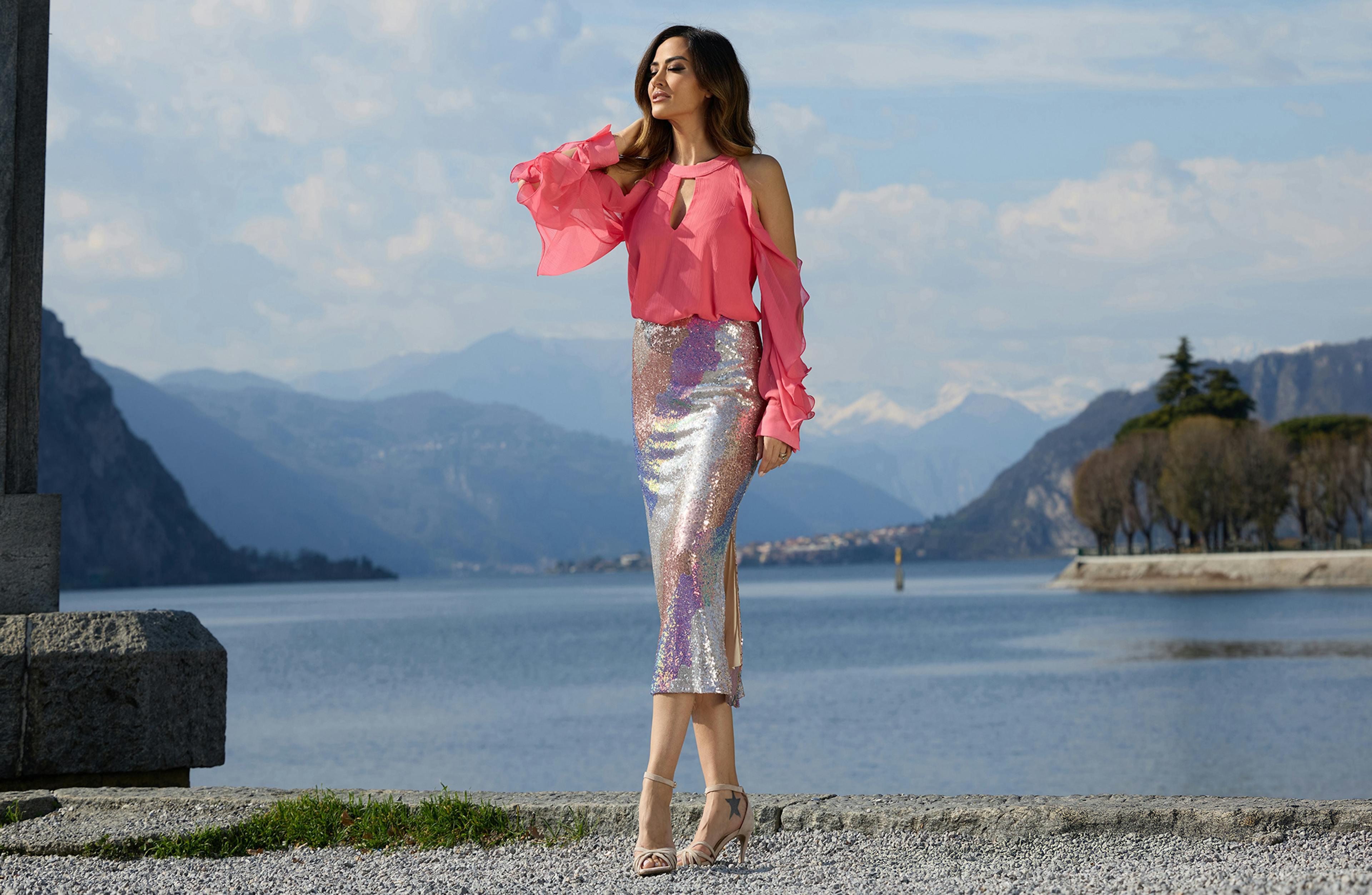 Giorgia Palmas in a pink blouse and shimmering skirt stands by a lake, with mountains in the background.