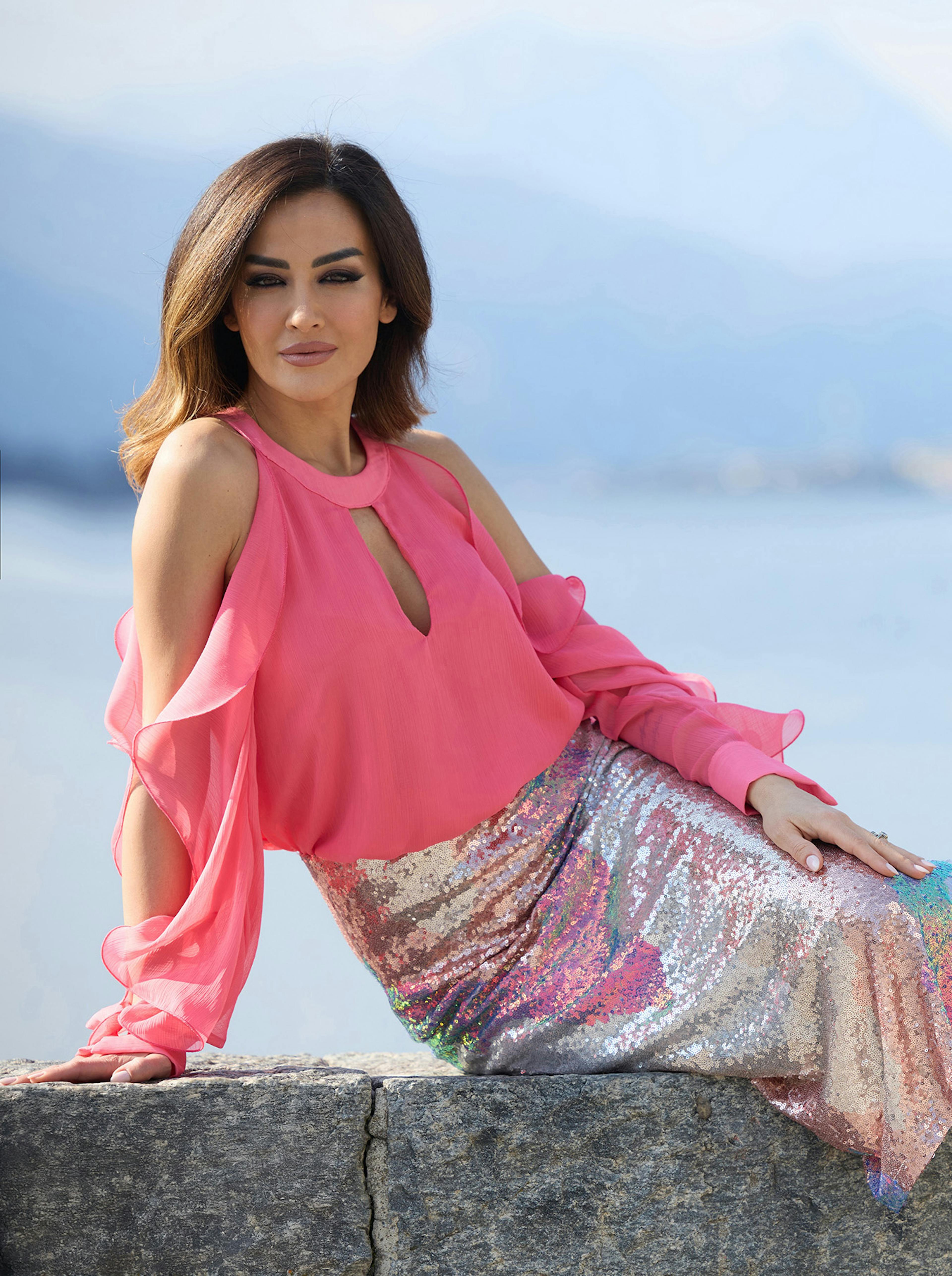 Giorgia Palmas in a bright pink top with flowing sleeves and a shimmering, sequin skirt sits confidently on a stone ledge by water.