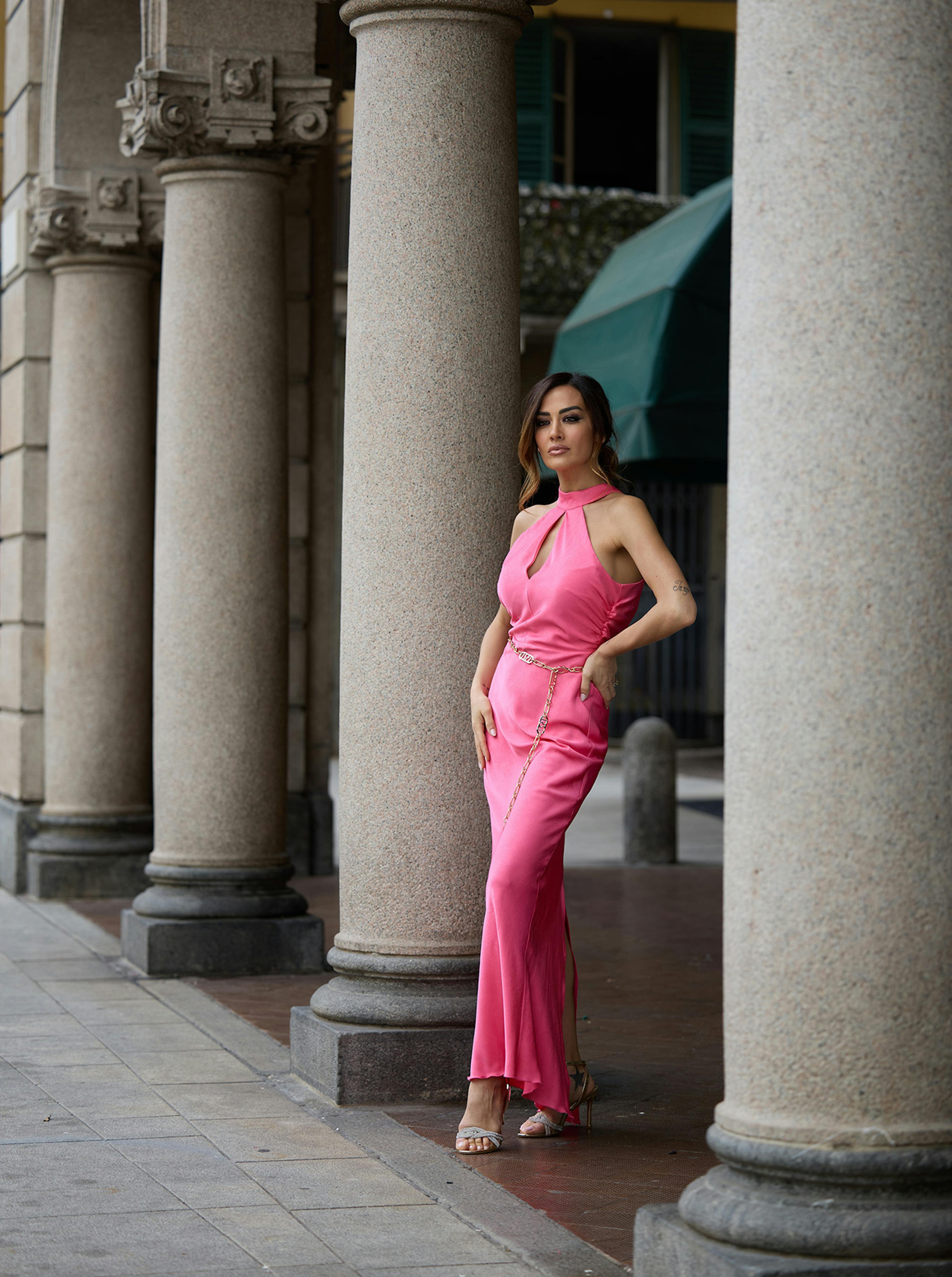 Giorgia Palmas in a pink dress stands confidently beside stone columns. The dress features a halter neck and gold chain accents, conveying elegance and grace.