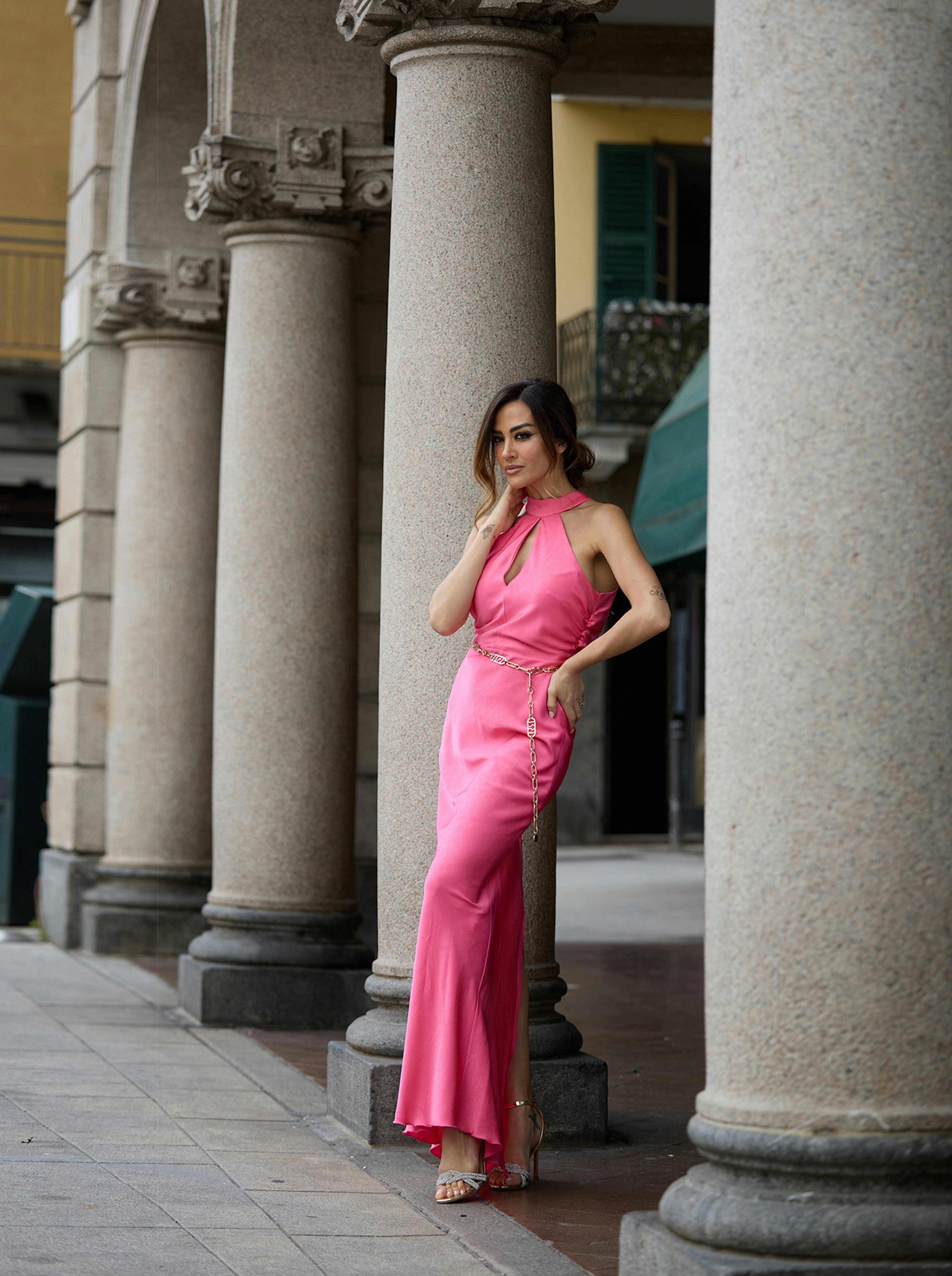 Giorgia Palmas in a pink dress stands confidently beside stone columns. The dress features a halter neck and gold chain accents, conveying elegance and grace.
