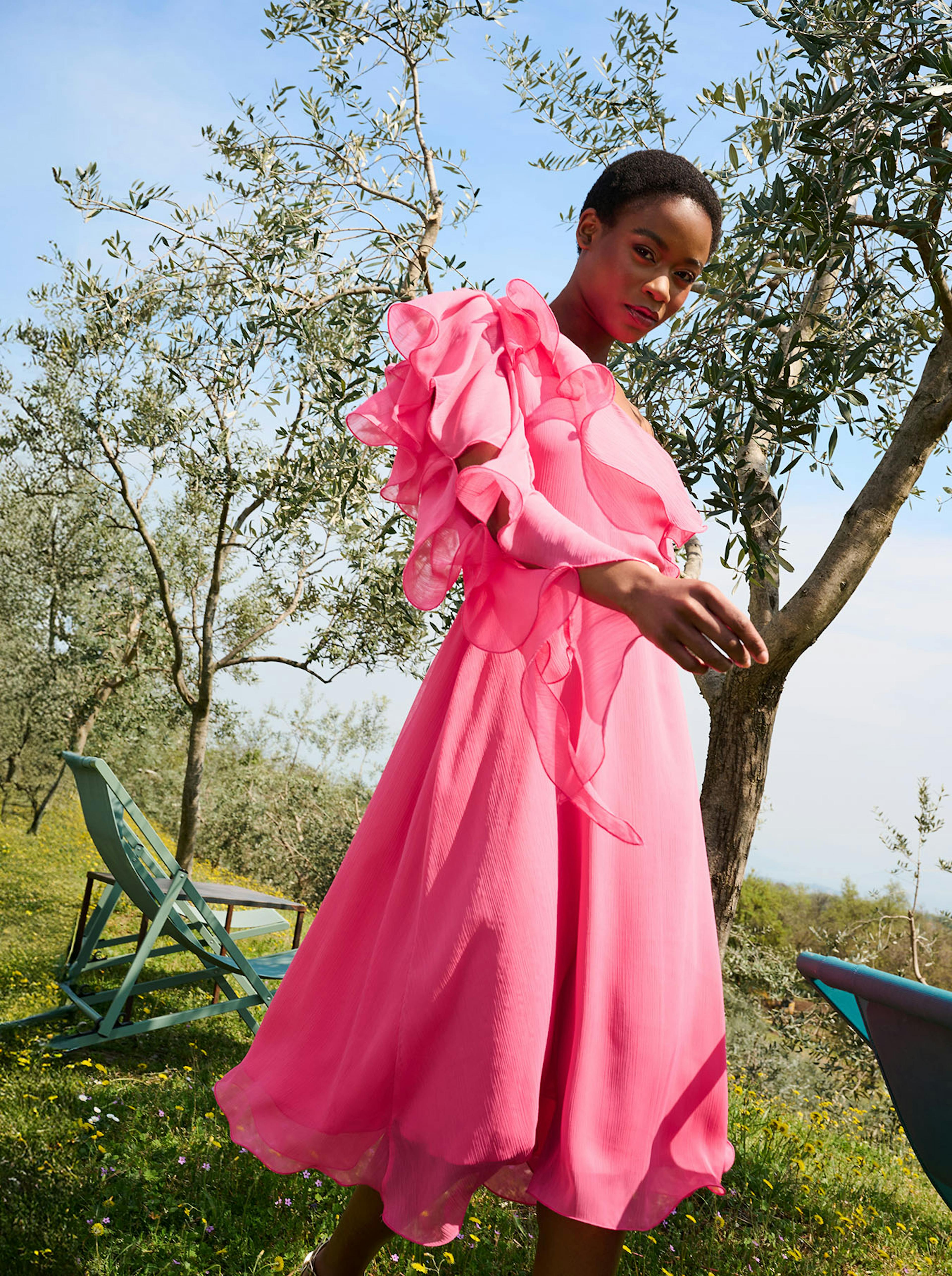 A woman in a flowing, vibrant pink dress with ruffled sleeves stands outdoors amid olive trees, exuding elegance and confidence under a clear sky.