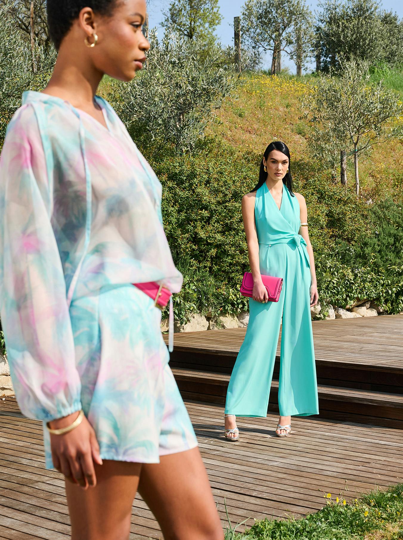 Two women in colorful summer outfits stand on a wooden deck surrounded by greenery. The foreground woman wears a pastel print dress; the background wears turquoise with a magenta clutch.