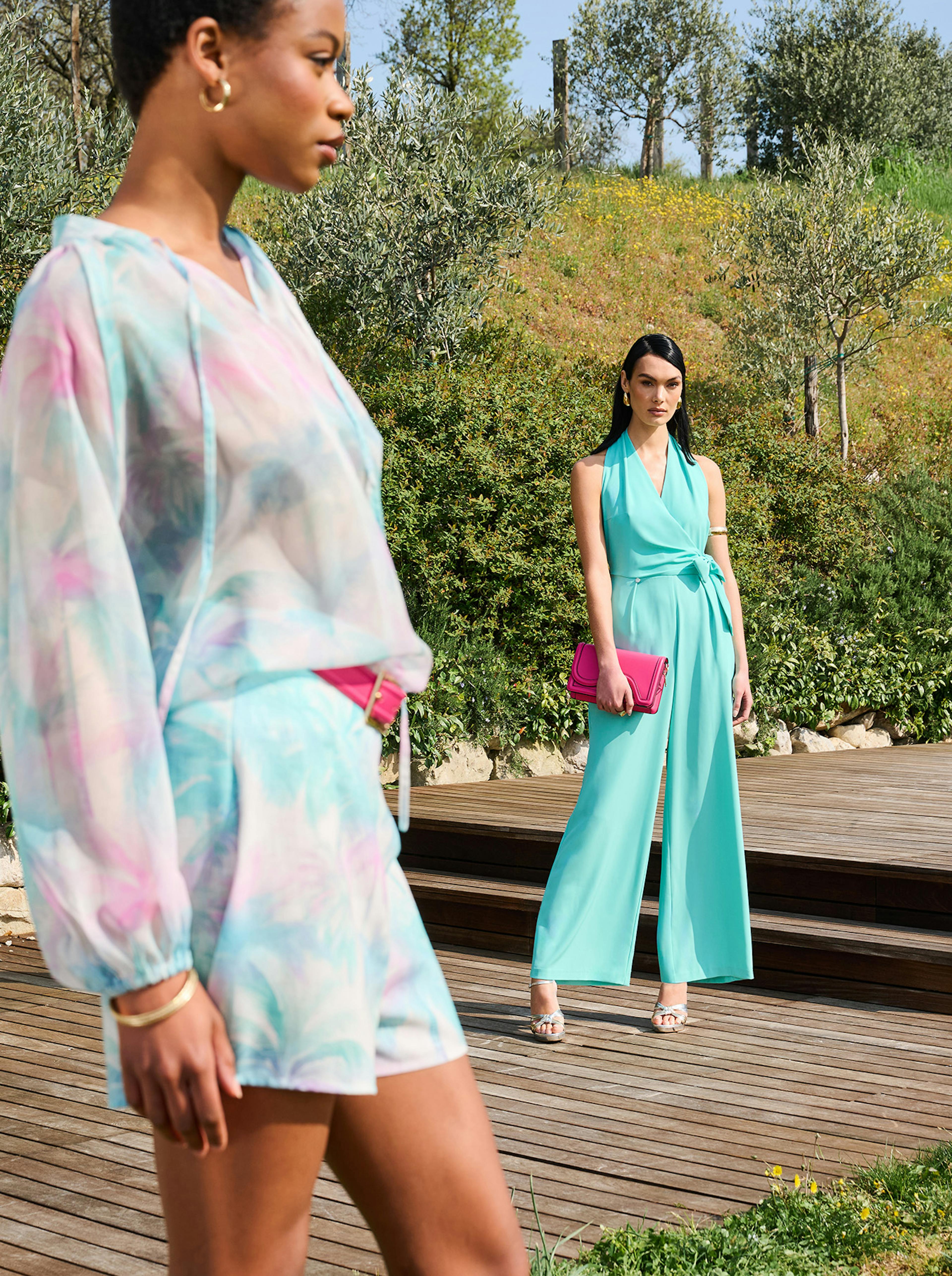 Two women in colorful summer outfits stand on a wooden deck surrounded by greenery. The foreground woman wears a pastel print dress; the background wears turquoise with a magenta clutch.