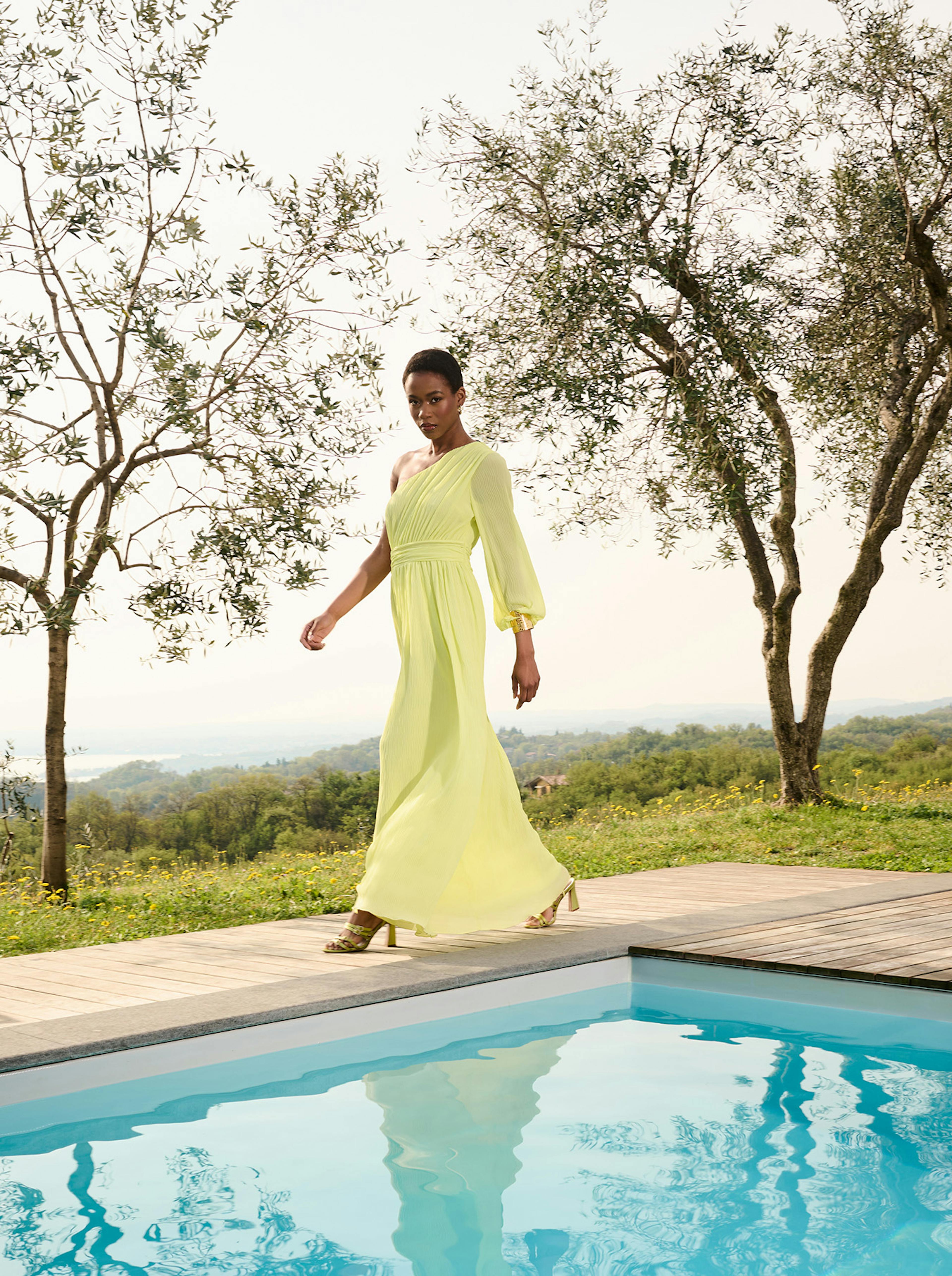 A woman in a flowing, one-shoulder yellow dress walks gracefully by a pool. Olive trees frame the scene, with a serene countryside in the background.
