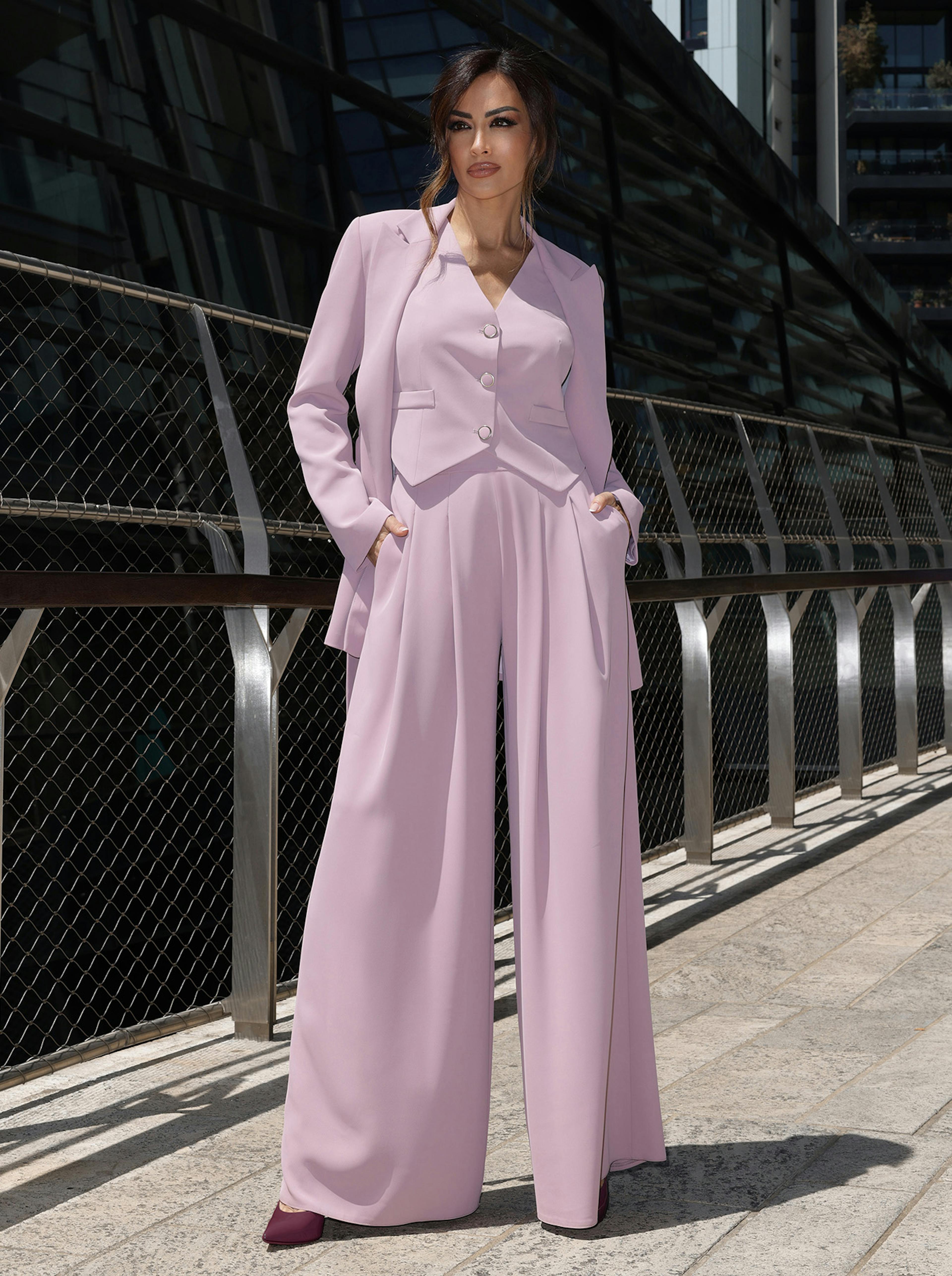 Giorgi Palmas stands confidently in front of a modern building, wearing a stylish lavender pantsuit. Her hands are in her pockets, exuding elegance and sophistication.