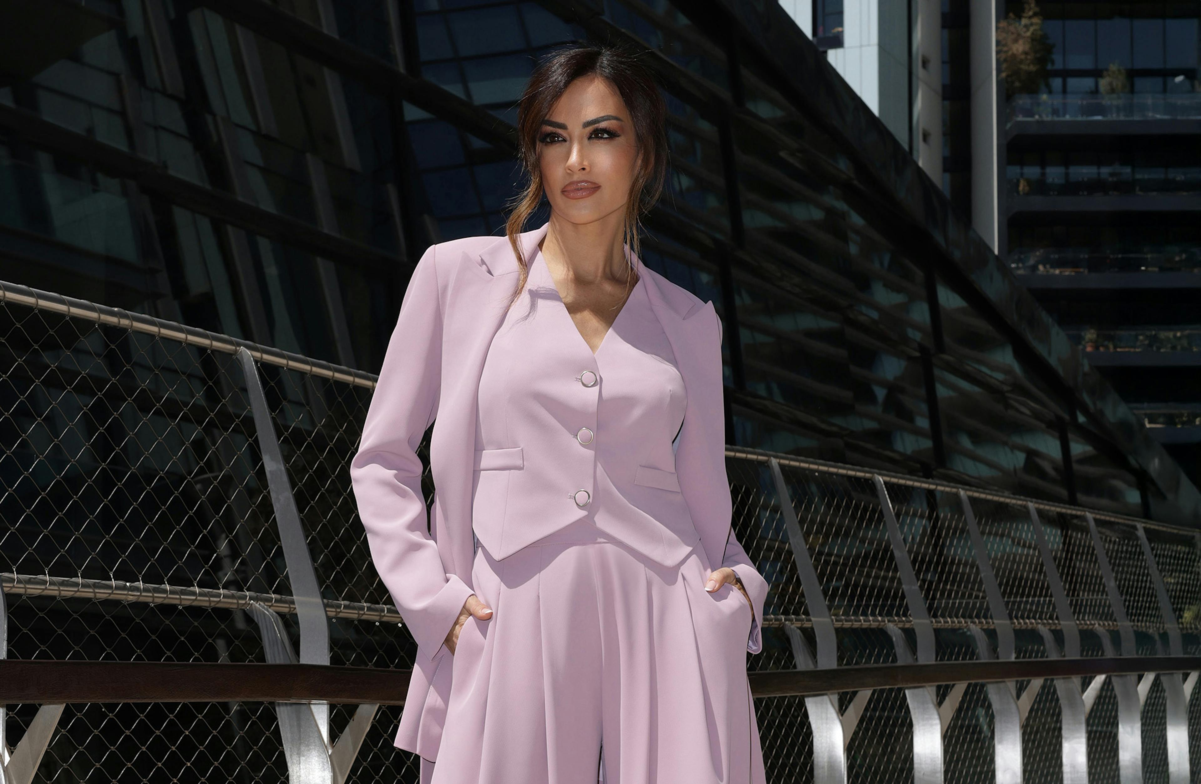 Giorgi Palmas stands confidently in front of a modern building, wearing a stylish lavender pantsuit. Her hands are in her pockets, exuding elegance and sophistication.