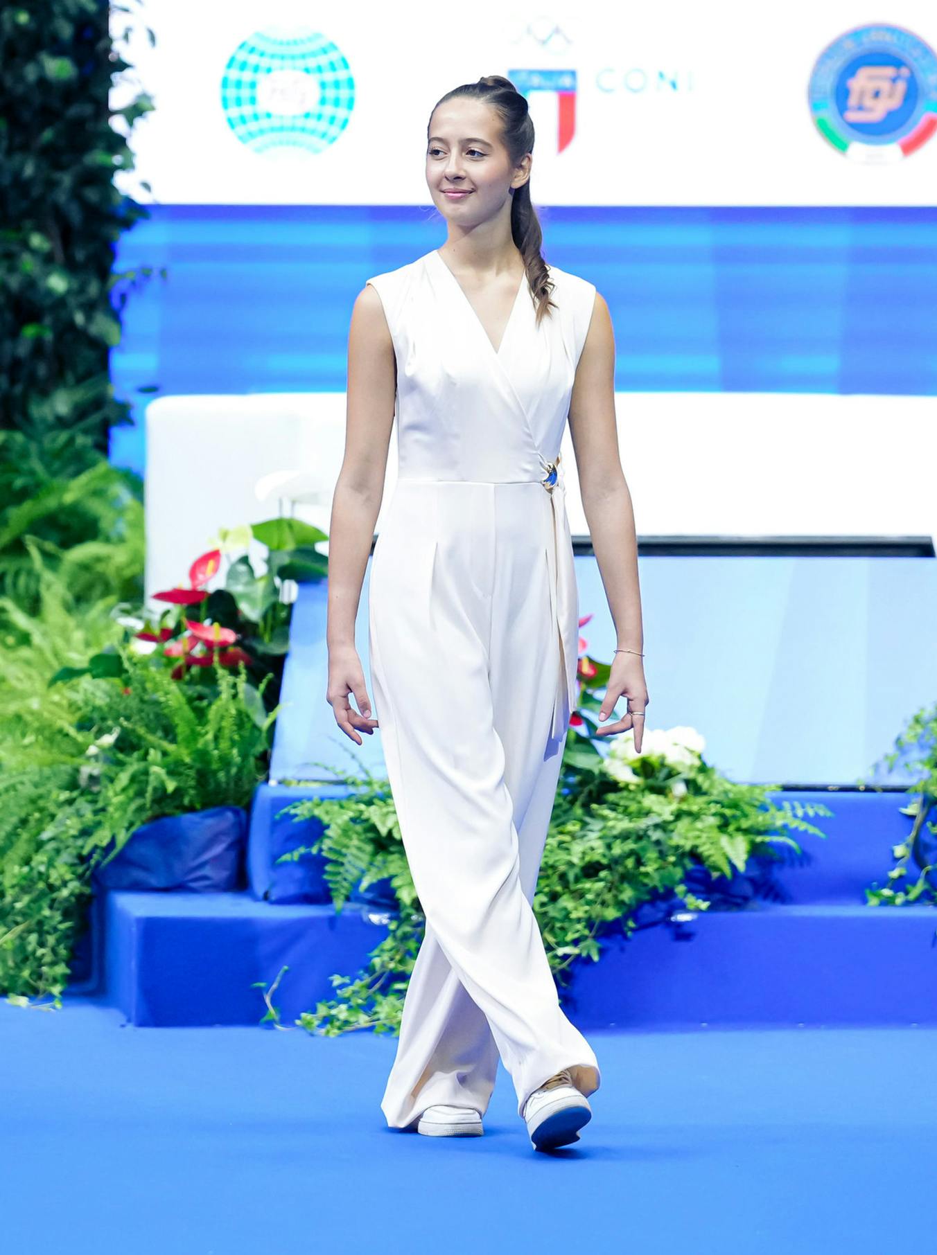 A smiling woman in a white jumpsuit stands on a blue stage, surrounded by green plants and colorful flowers, exuding elegance and confidence.
