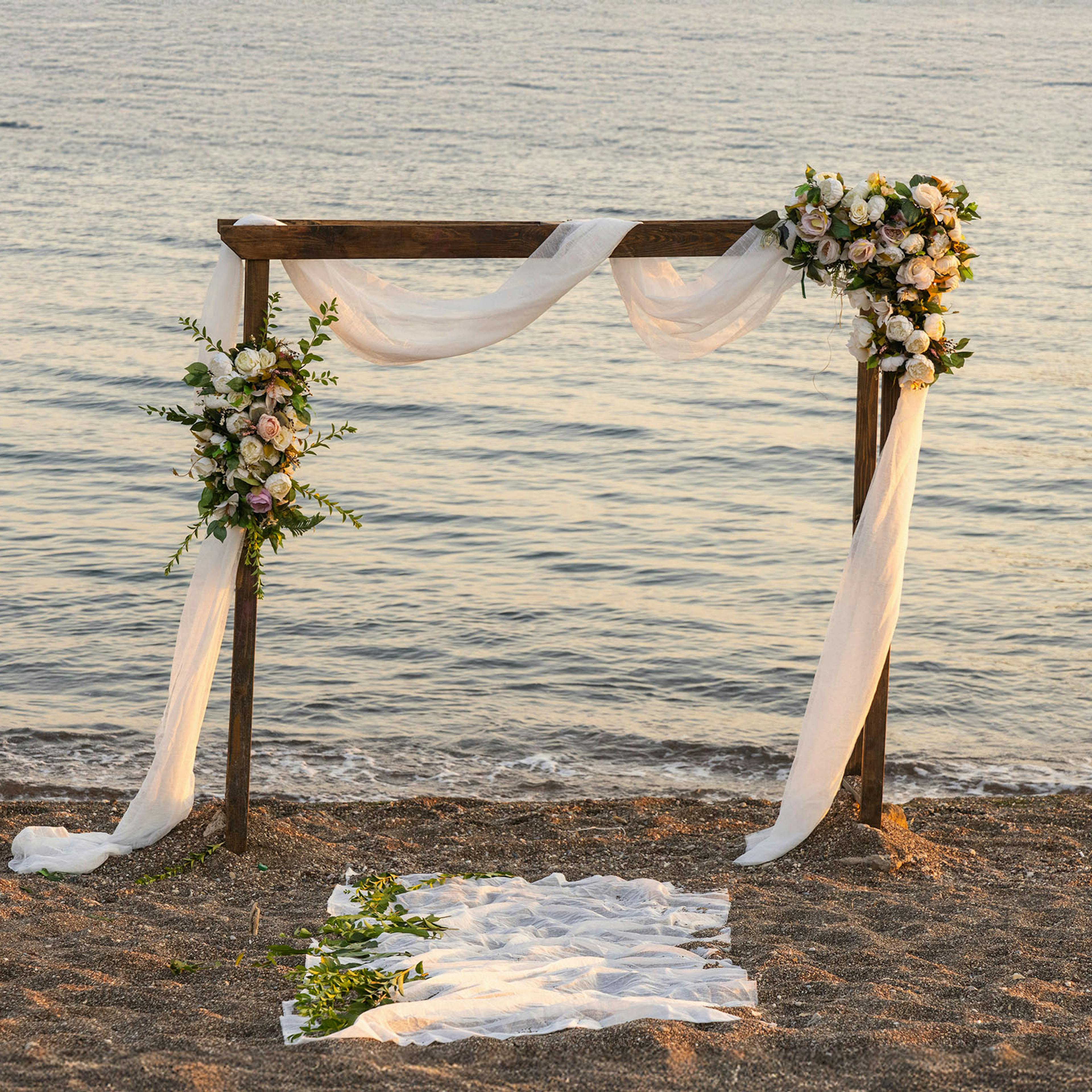 A wooden wedding arch draped in white fabric and adorned with white and pink flowers stands on a sandy beach.