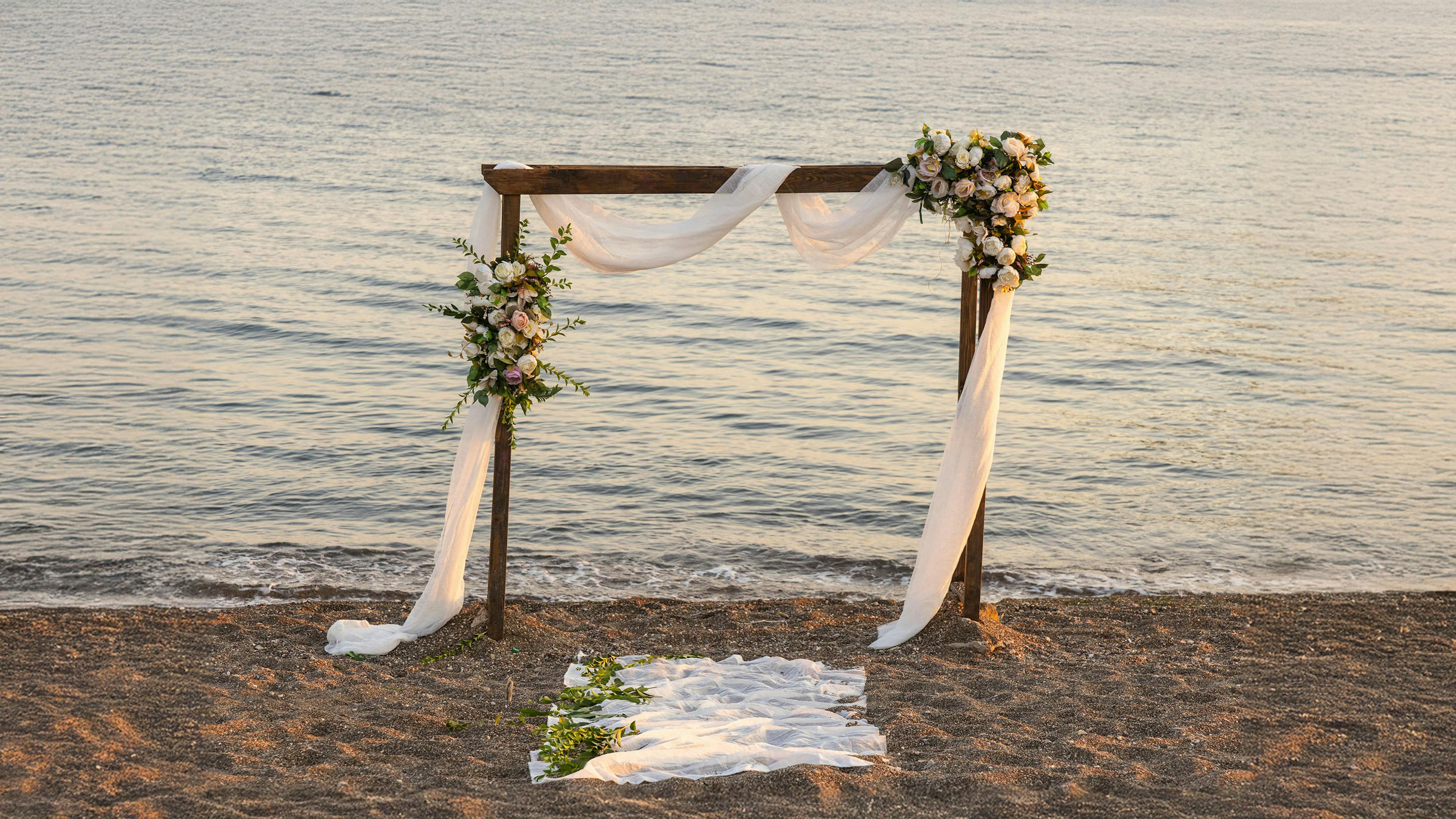A wooden wedding arch draped in white fabric and adorned with white and pink flowers stands on a sandy beach.