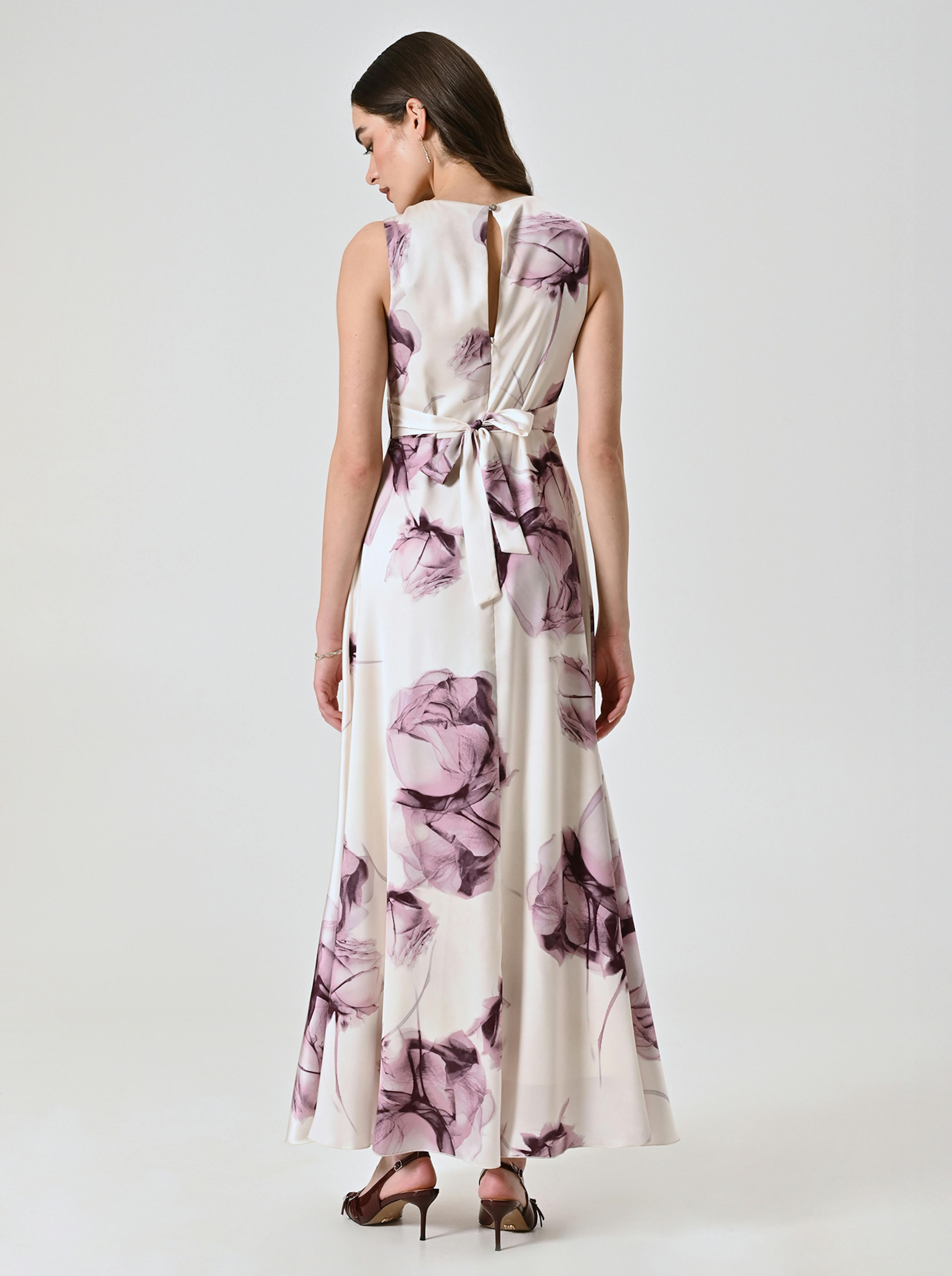 Woman in a sleeveless, long white dress with purple floral prints, tied at the waist, standing against a plain background. The mood is elegant.