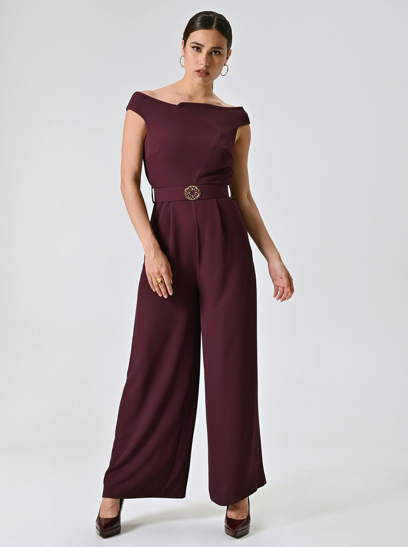 Woman in an elegant maroon jumpsuit with an off-shoulder design and wide legs. She wears hoop earrings and heels, exuding confidence against a plain backdrop.