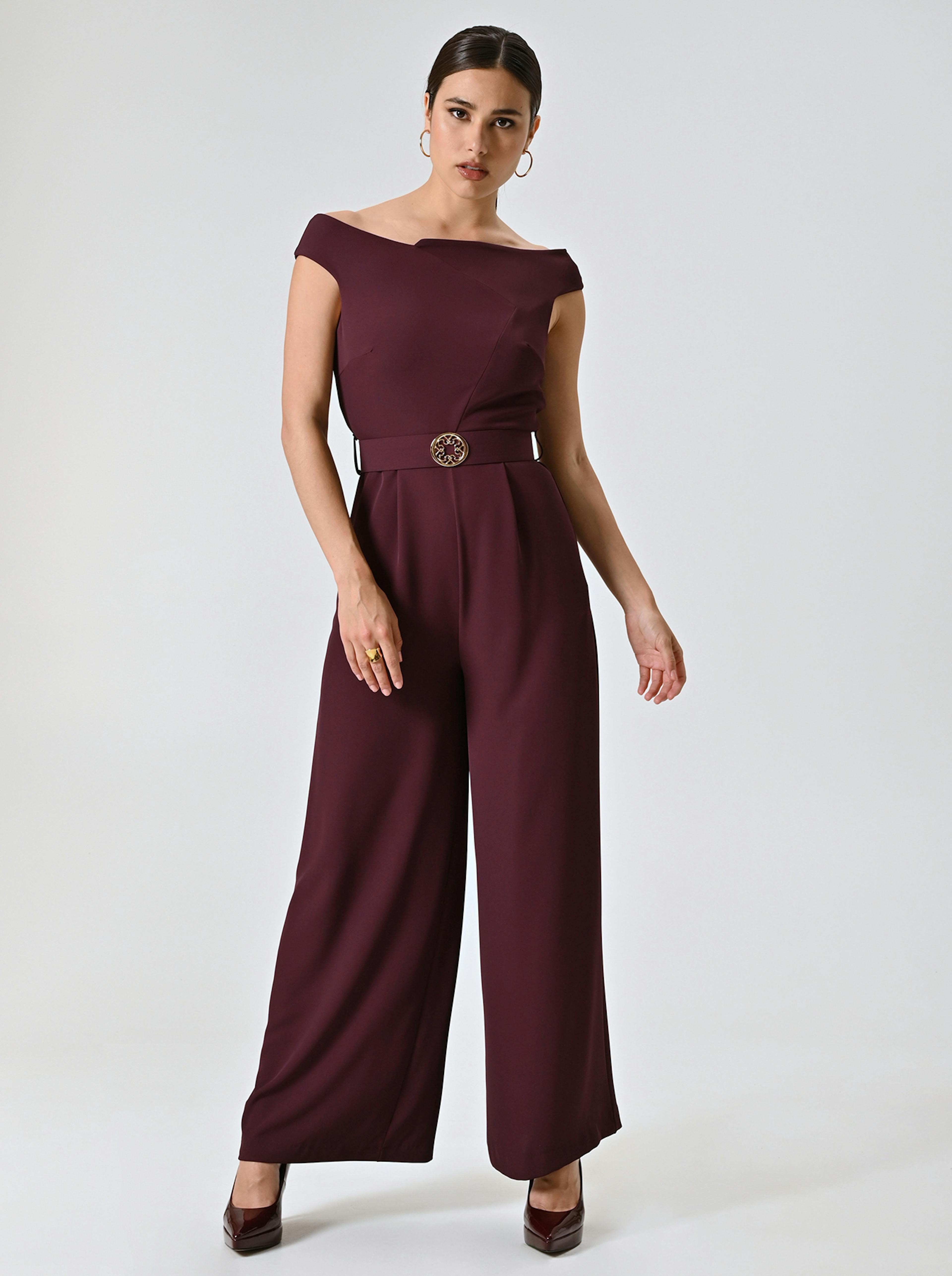 Woman in an elegant maroon jumpsuit with an off-shoulder design and wide legs. She wears hoop earrings and heels, exuding confidence against a plain backdrop.